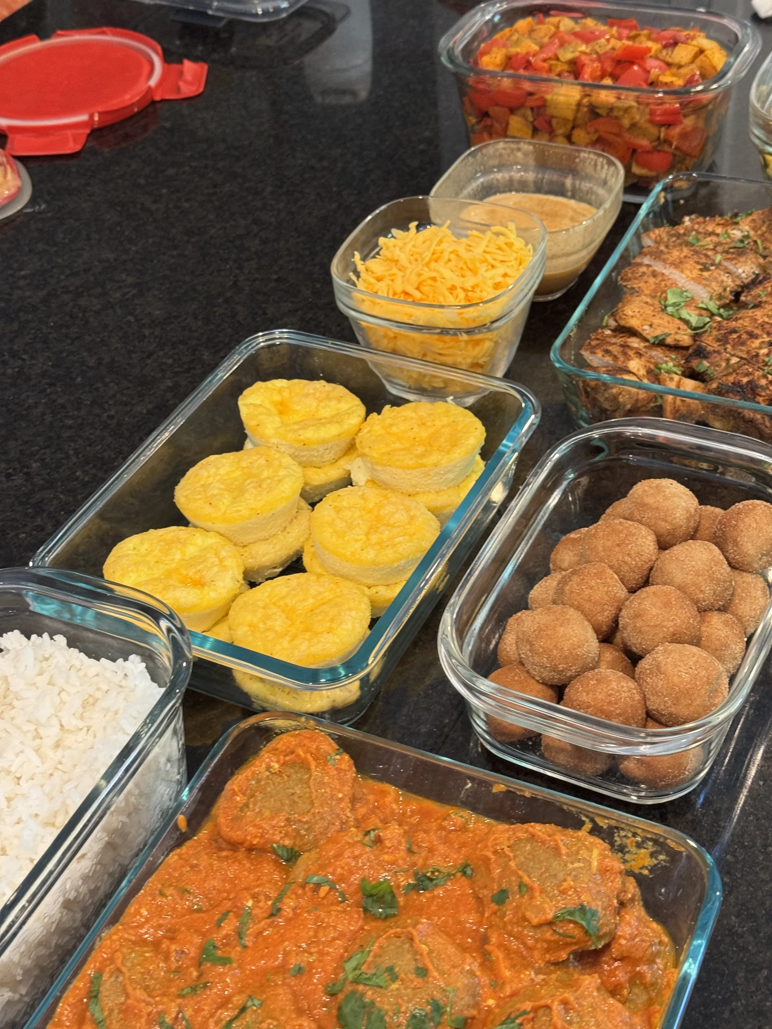 Assorted dishes in glass containers on a black countertop, including yellow corn muffins, shredded cheese, cooked vegetables, fried meat, and a bowl of white rice.