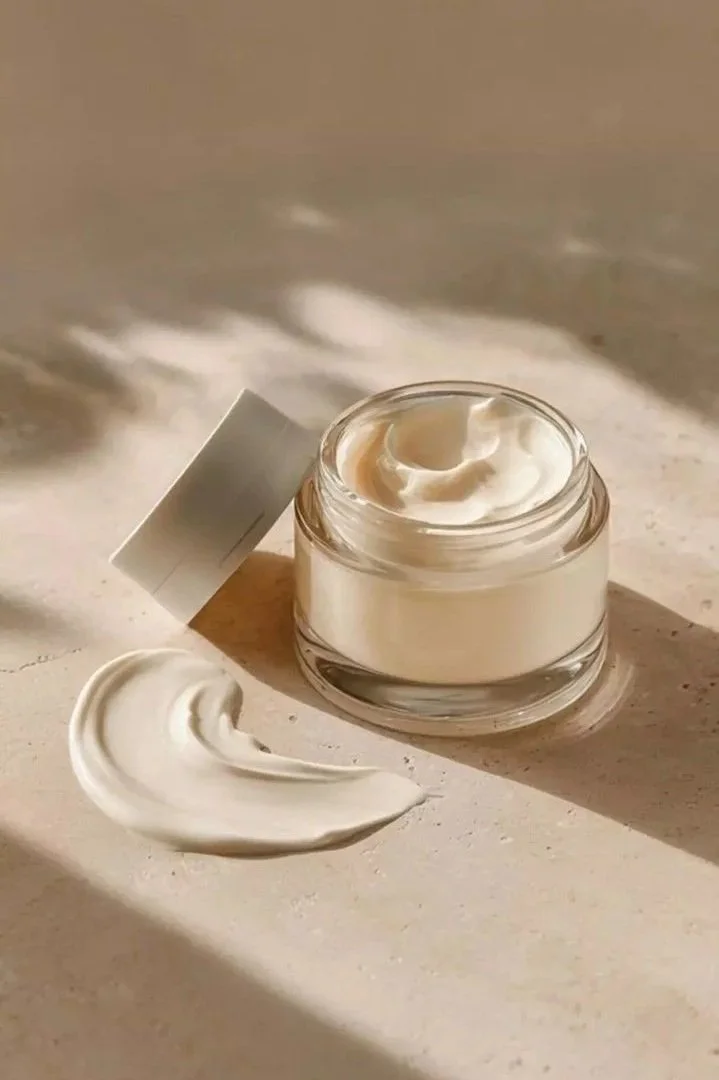 Crème hydratante dans un petit pot en verre avec le couvercle posé à côté. Une quantité de crème est étalée sur une surface claire, avec un fond neutre lumineux.