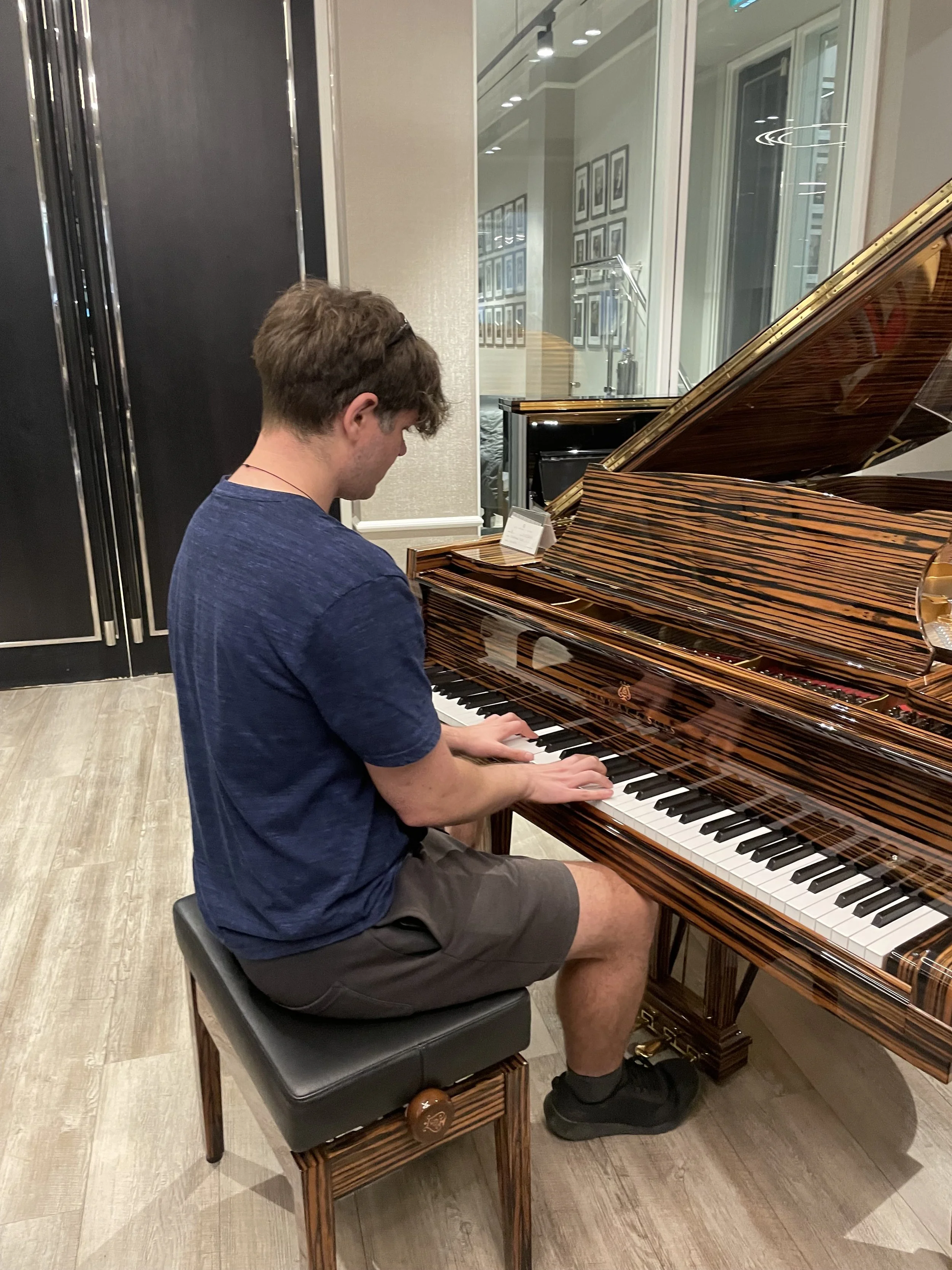 A young man sitting on a piano bench, playing a grand piano in a room with wall art and glass windows.