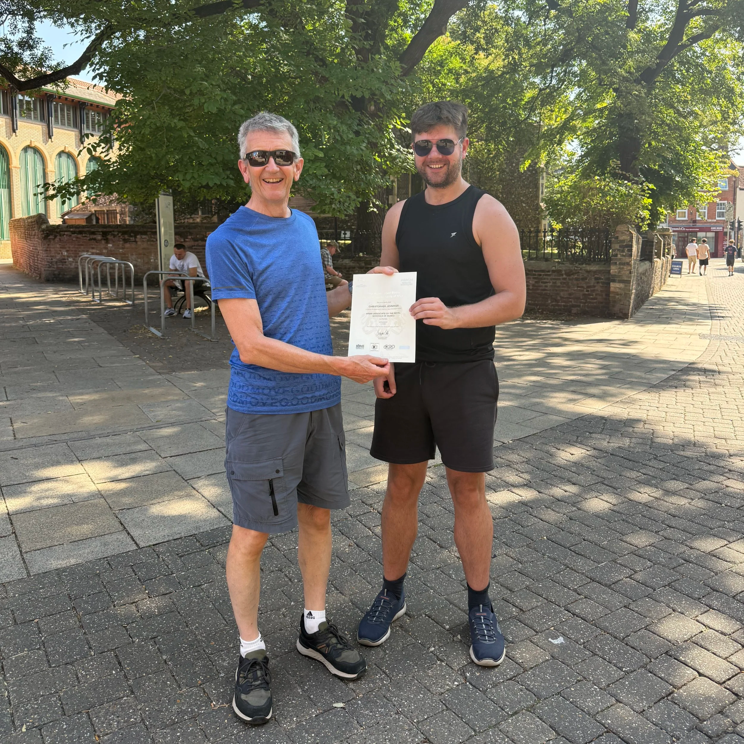 Two men are standing outdoors under a large leafy tree, smiling and holding a certificate. One man is older with gray hair, wearing sunglasses, a blue shirt, and gray shorts. The other man is younger with dark hair and a beard, wearing sunglasses, a black sleeveless shirt, black shorts, and sneakers. In the background, there are a few people sitting on benches and walking, with brick buildings and a church open sign visible.