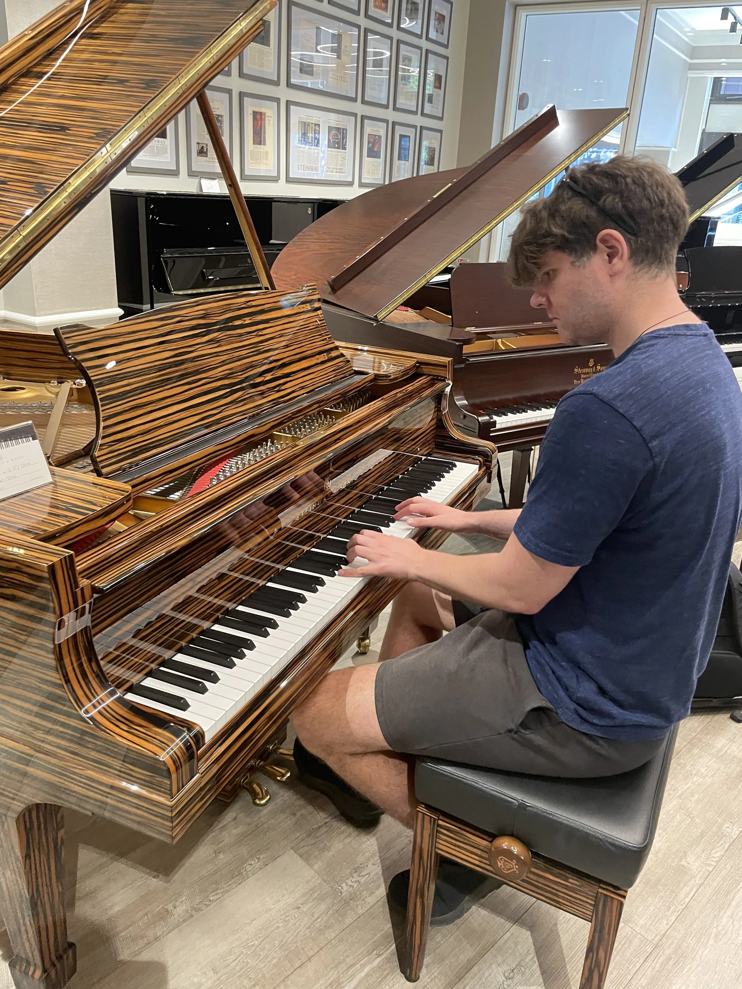 Man playing a grand piano with a tiger-stripe wood finish in a room with framed pictures on the wall.