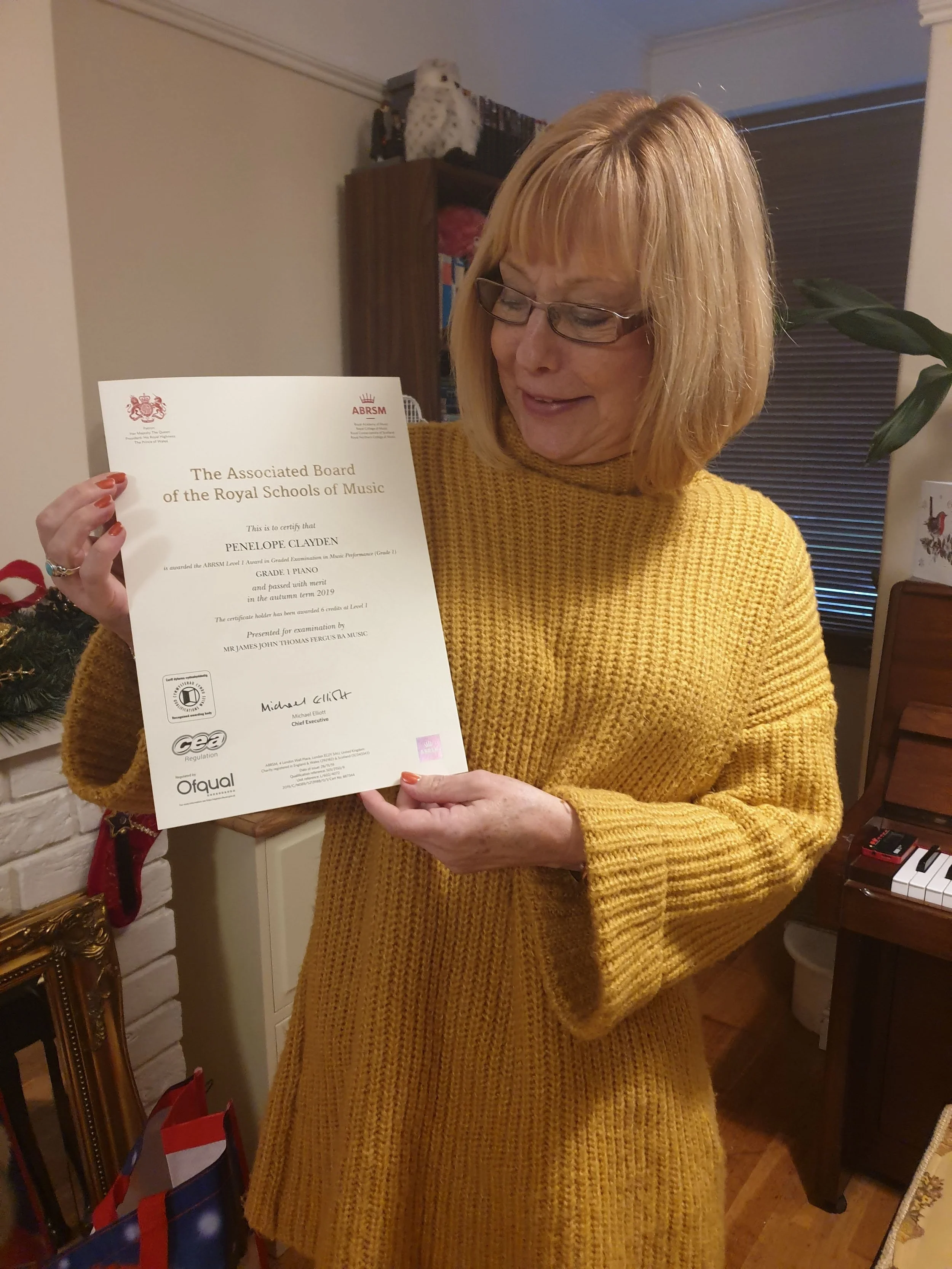 A woman with blonde hair and glasses in a yellow sweater smiling while holding a certificate of achievement. There are gift bags and holiday decorations in the background.