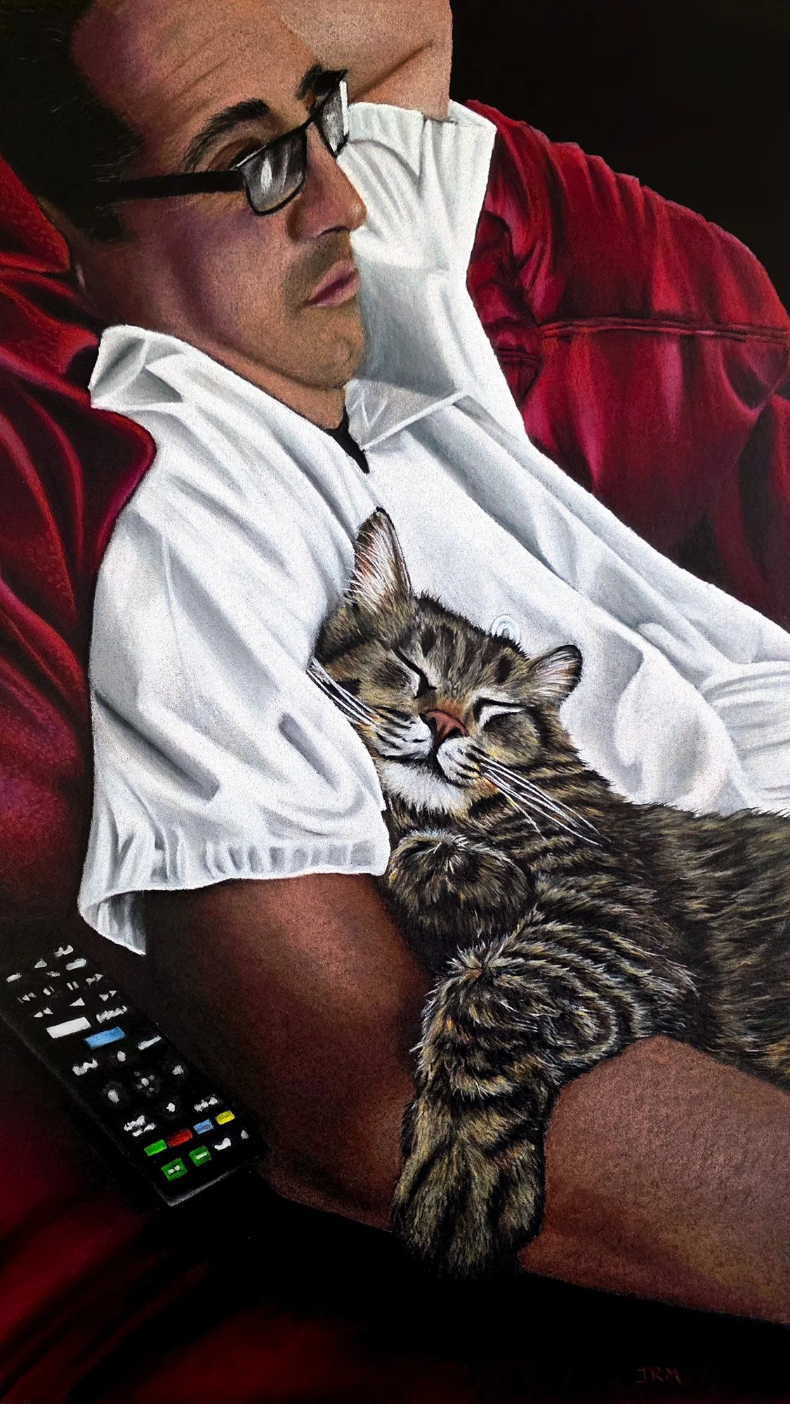 A man with glasses and short hair is resting on a red blanket, holding a sleeping tabby kitten with closed eyes and relaxed posture.
