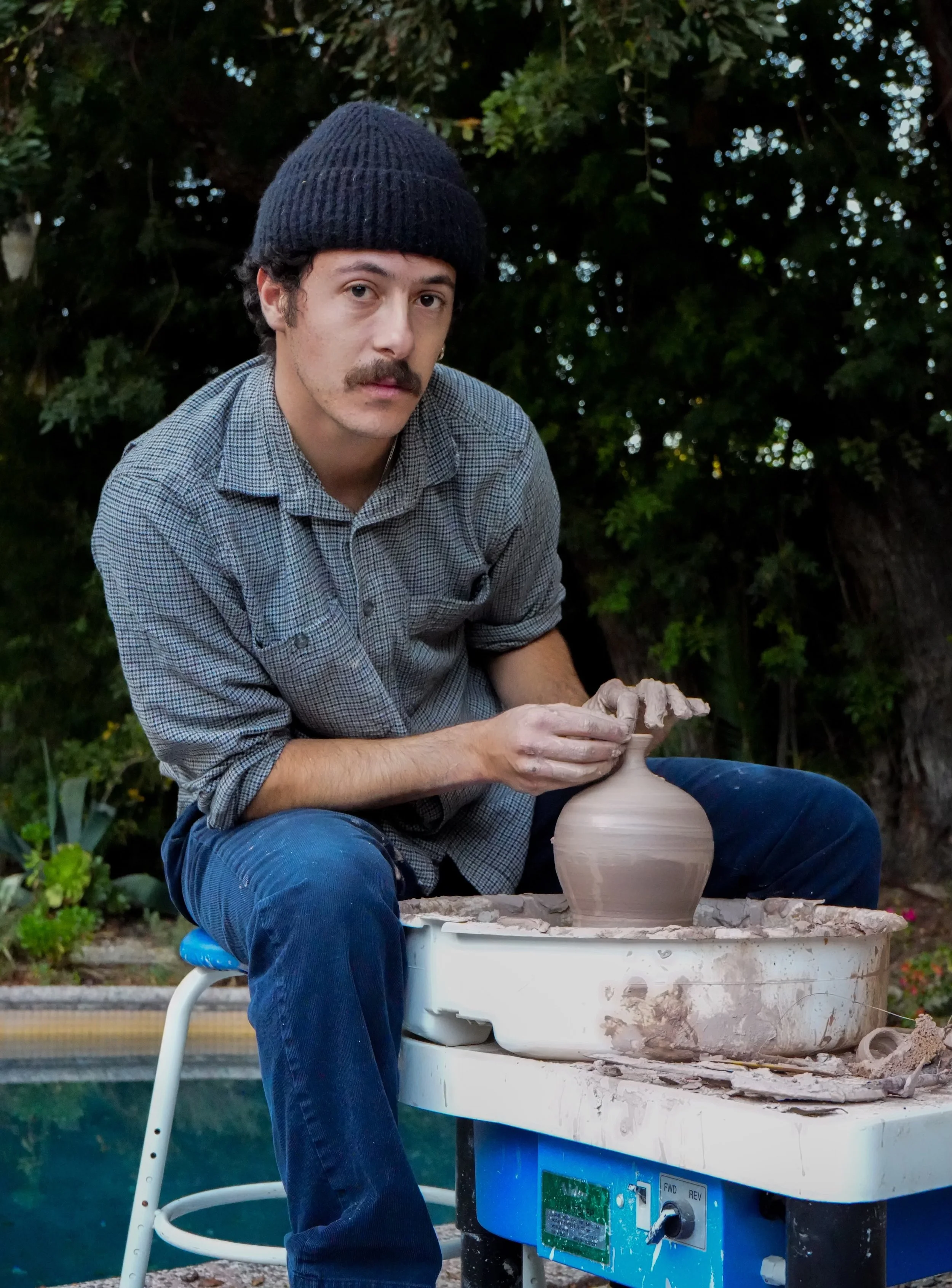 A man with a dark knit cap, checked shirt, and jeans sits outdoors at a pottery wheel shaping a clay vase during daytime.