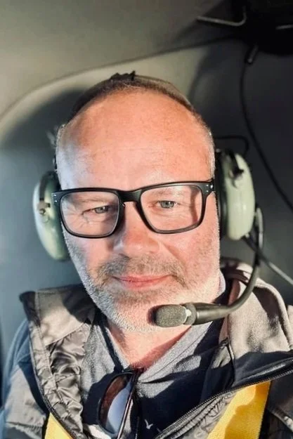 A man wearing glasses, a headset with a microphone, and a jacket, sitting inside the cockpit of an aircraft.