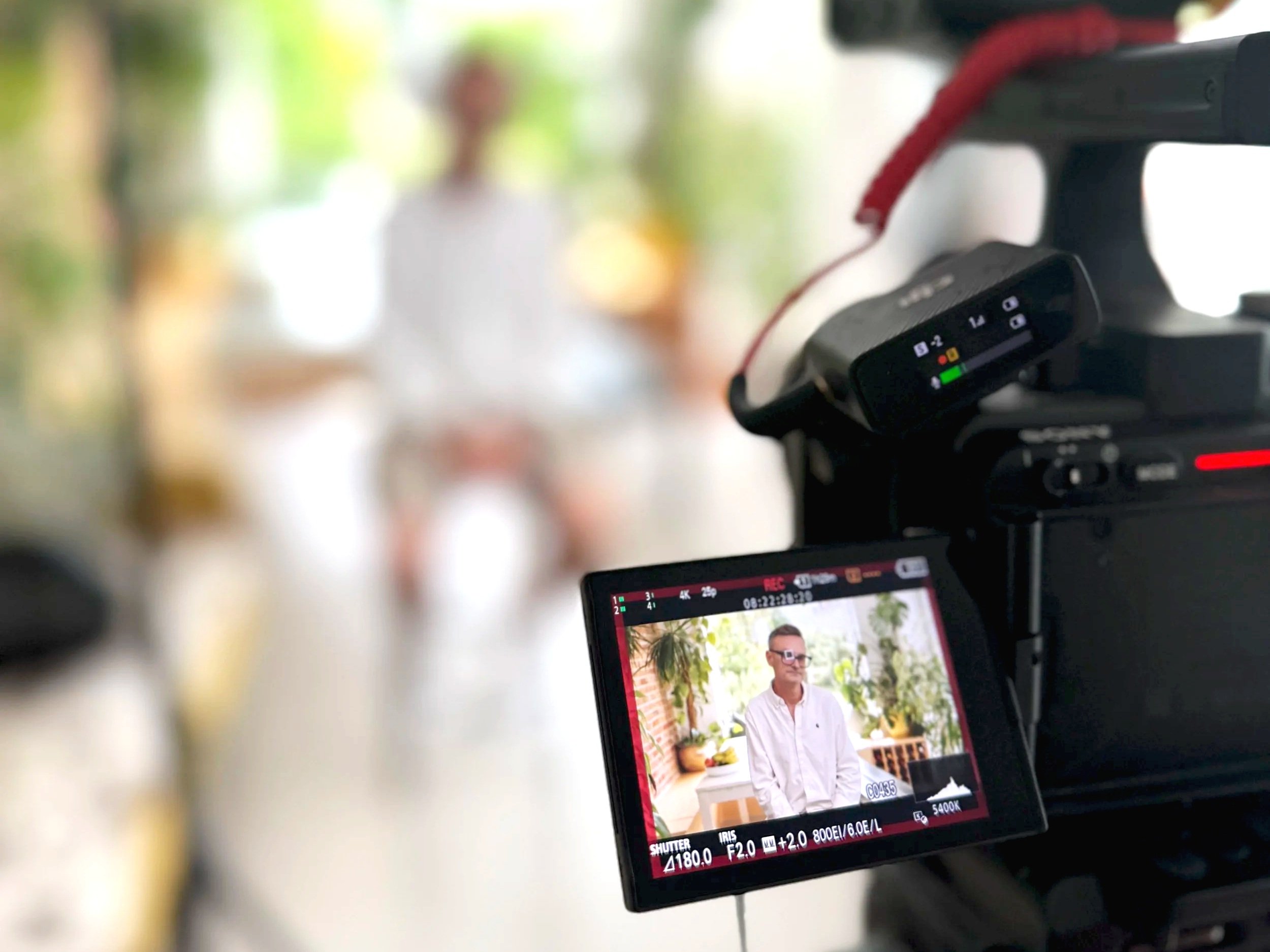 A professional video camera with a monitor displaying a man in glasses and a white shirt, set in a bright indoor space with blurred background of greenery.