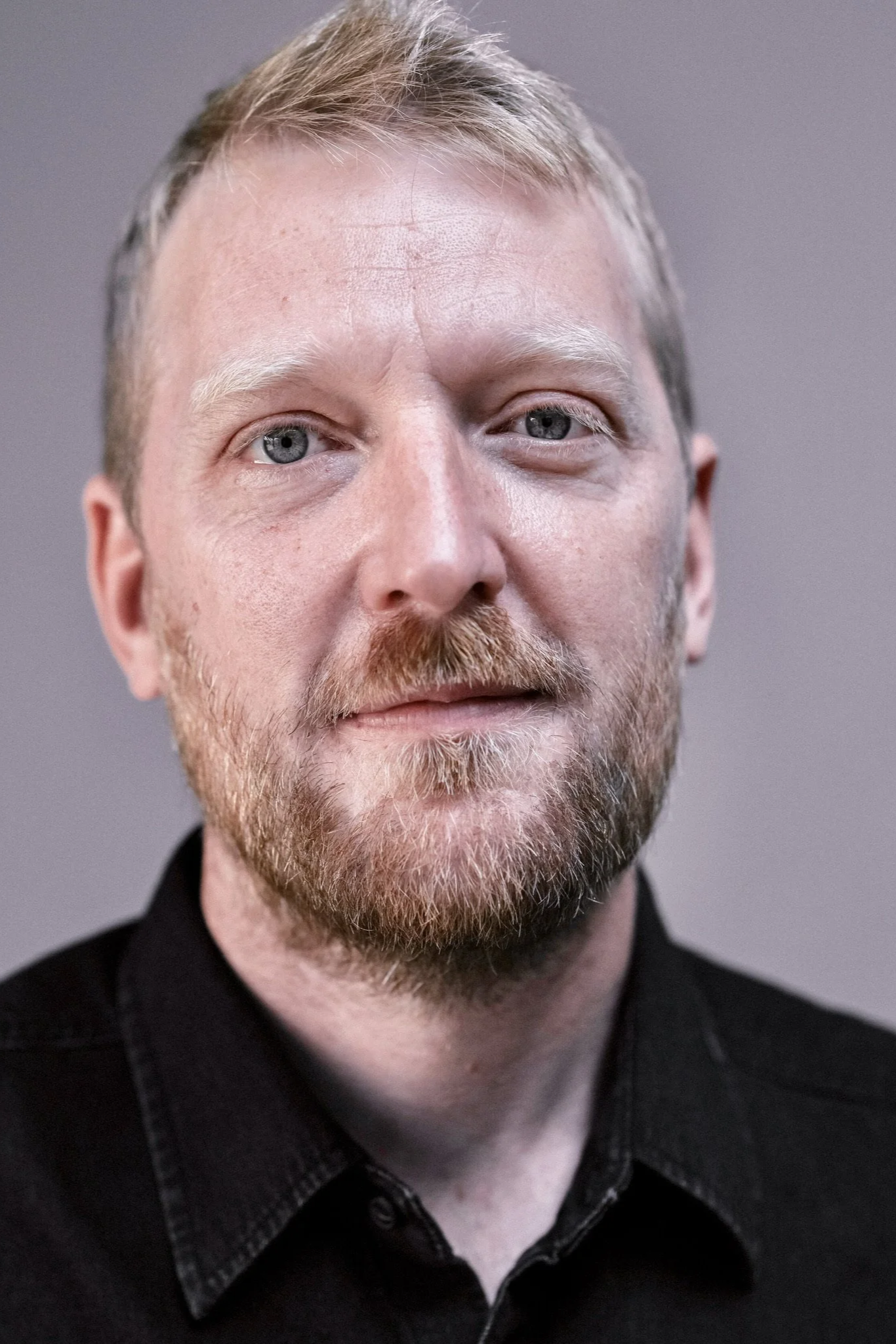 Close-up of a Caucasian man with short blond hair, blue eyes, and a beard, wearing a black collared shirt, looking directly at the camera against a plain gray background.