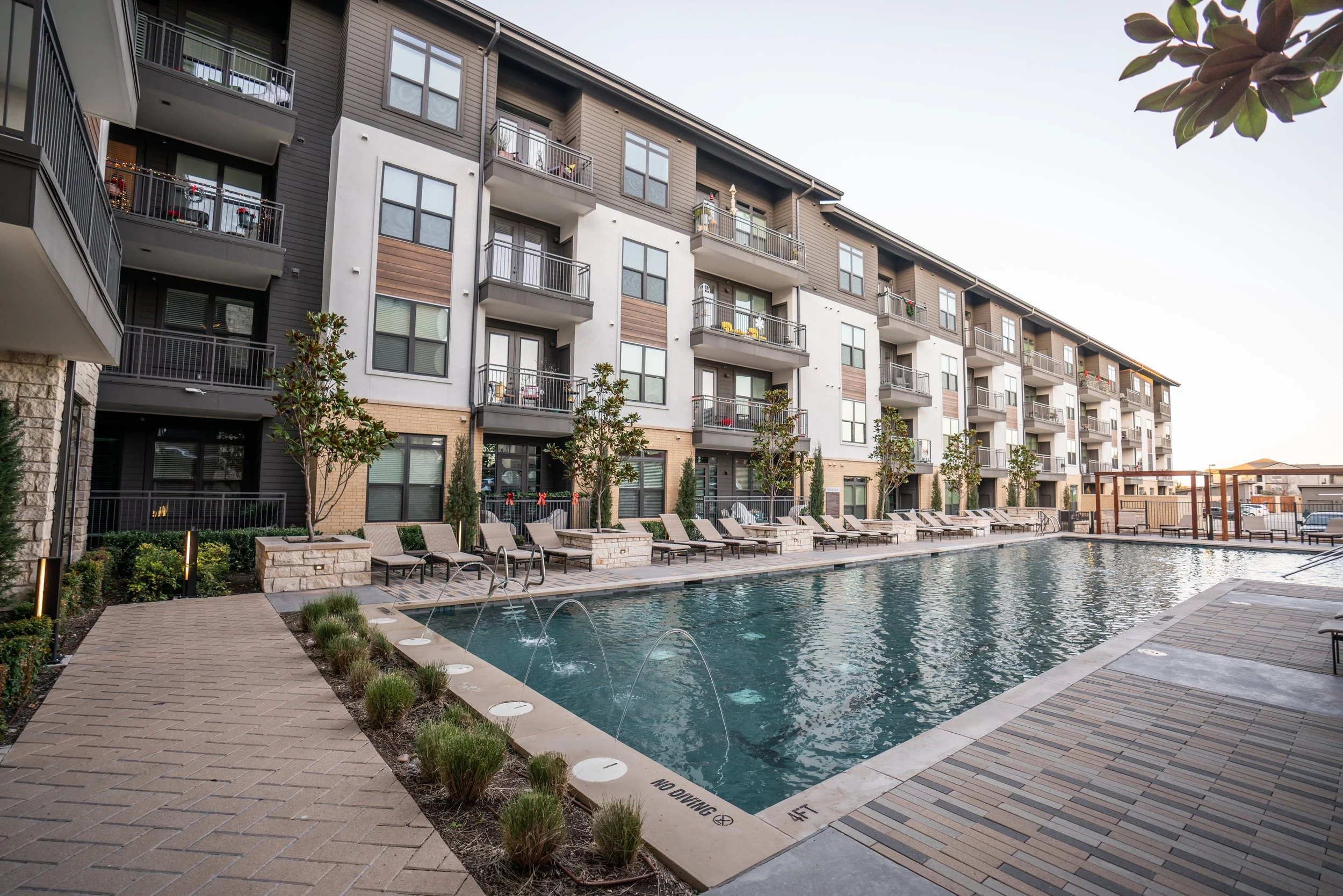 Apartment building with multiple balconies overlooking a swimming pool surrounded by lounge chairs and small landscaped trees.