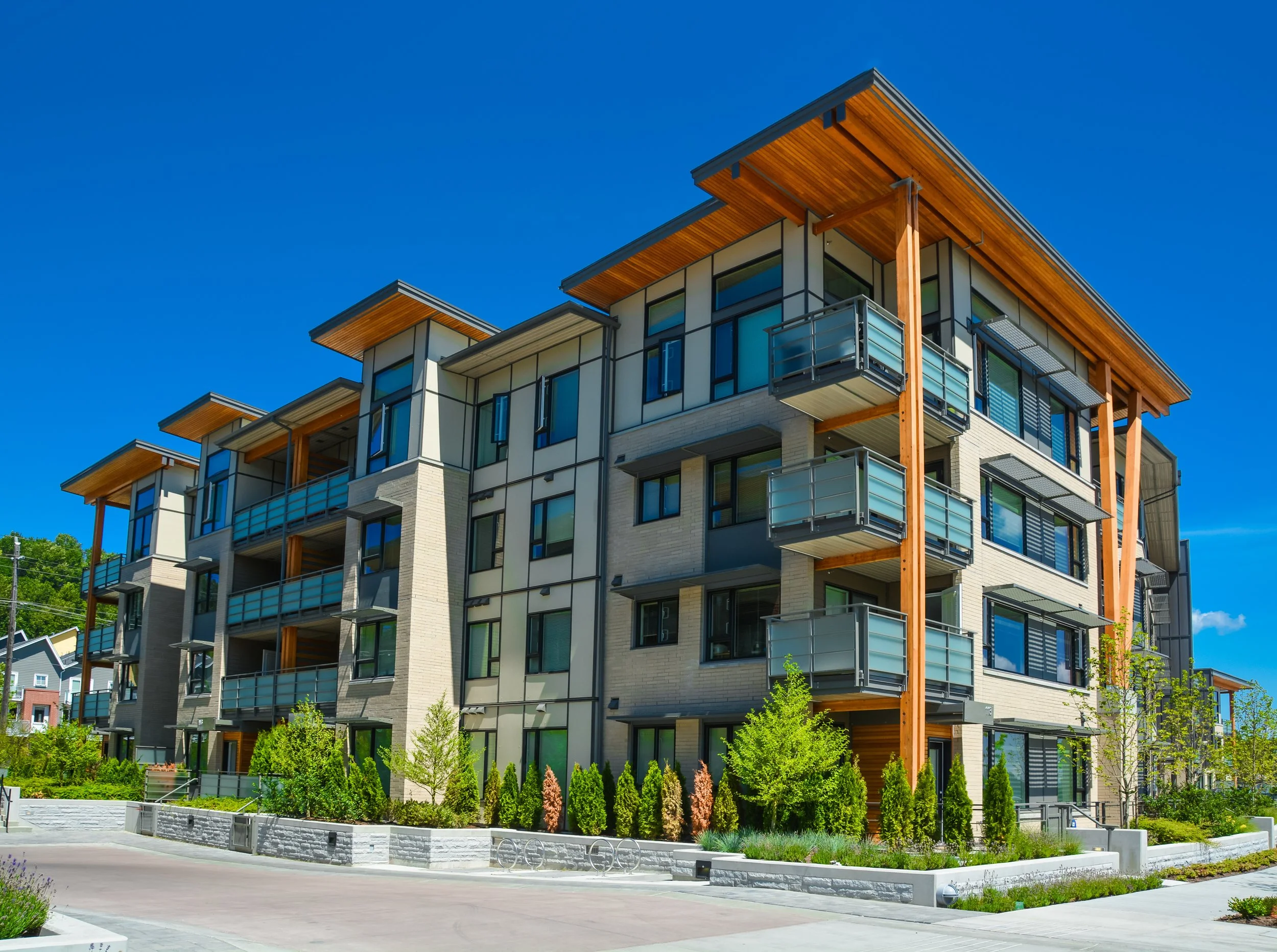 Modern multi-story apartment building with balconies, wooden accents, large windows, and landscaped front yard against a bright blue sky.