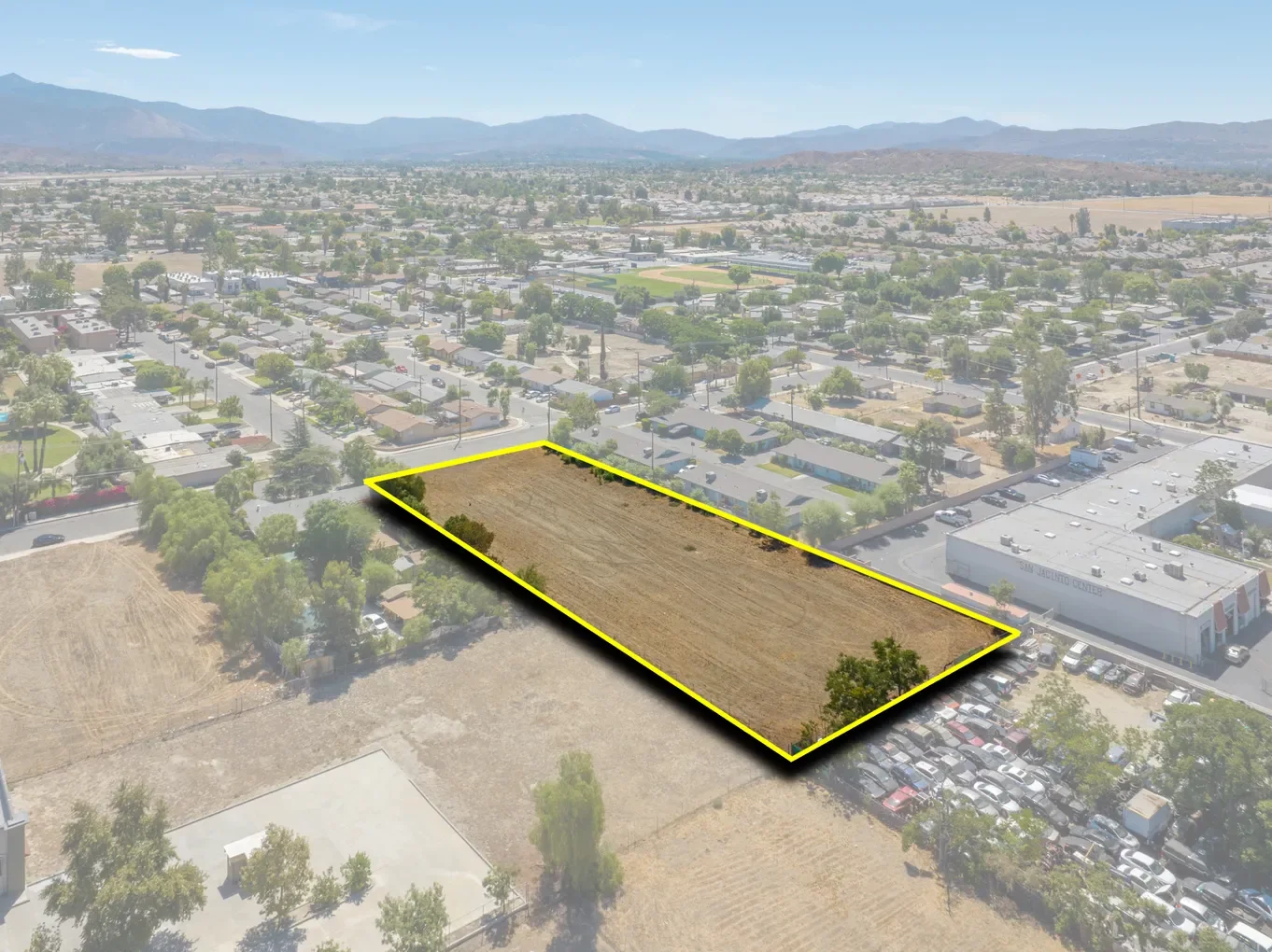 Aerial view of a vacant lot outlined in yellow in an urban area with businesses, parking lots, and residential homes, with mountains in the background.