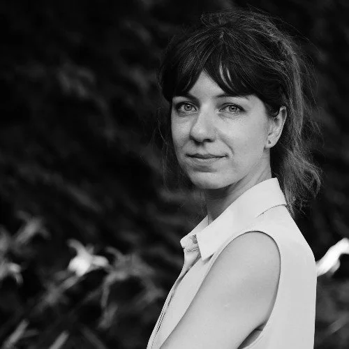 Black and white photo of a woman with dark hair, wearing a sleeveless top, standing outdoors with blurred foliage in the background.