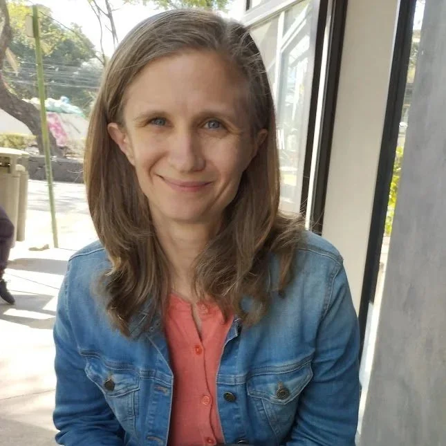 A woman with shoulder-length brown hair, blue eyes, and fair skin sitting in a well-lit indoor space near large windows, wearing a denim jacket over a pink shirt, smiling softly.