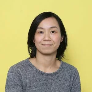 Portrait of a woman with black hair wearing a gray shirt against a yellow background.