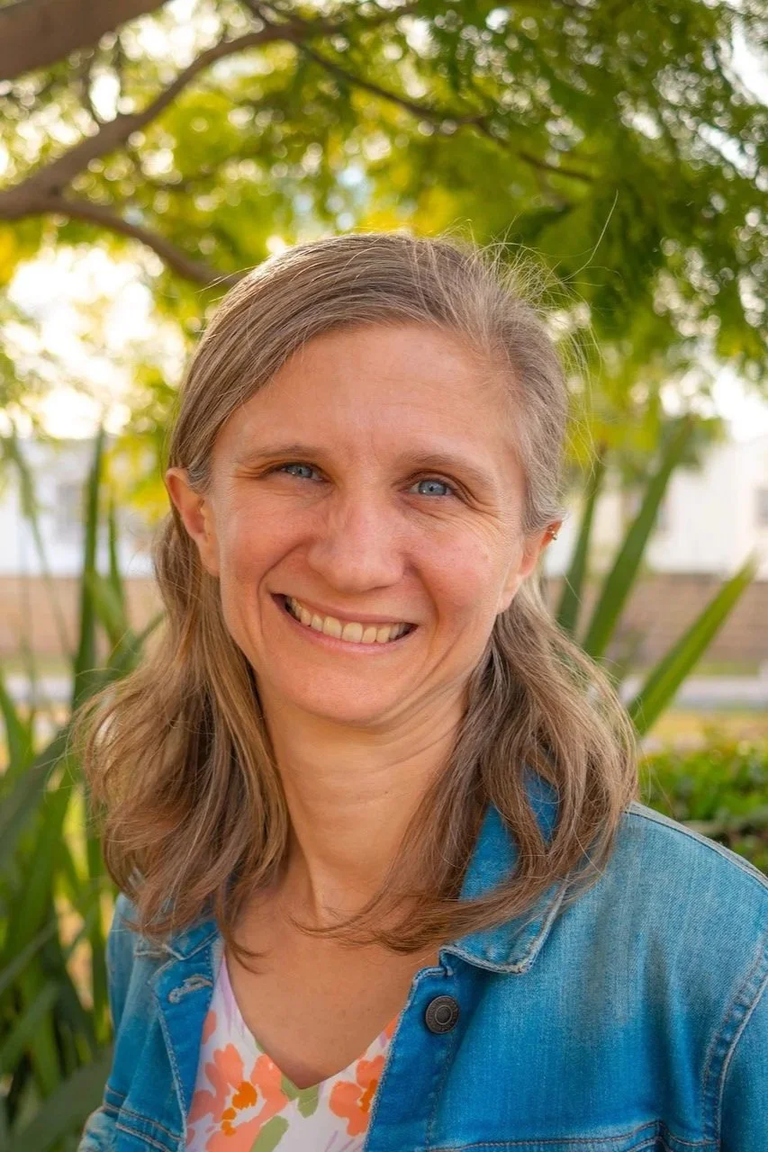 A woman smiling outdoors with trees and green foliage in the background, wearing a denim jacket over a floral top.