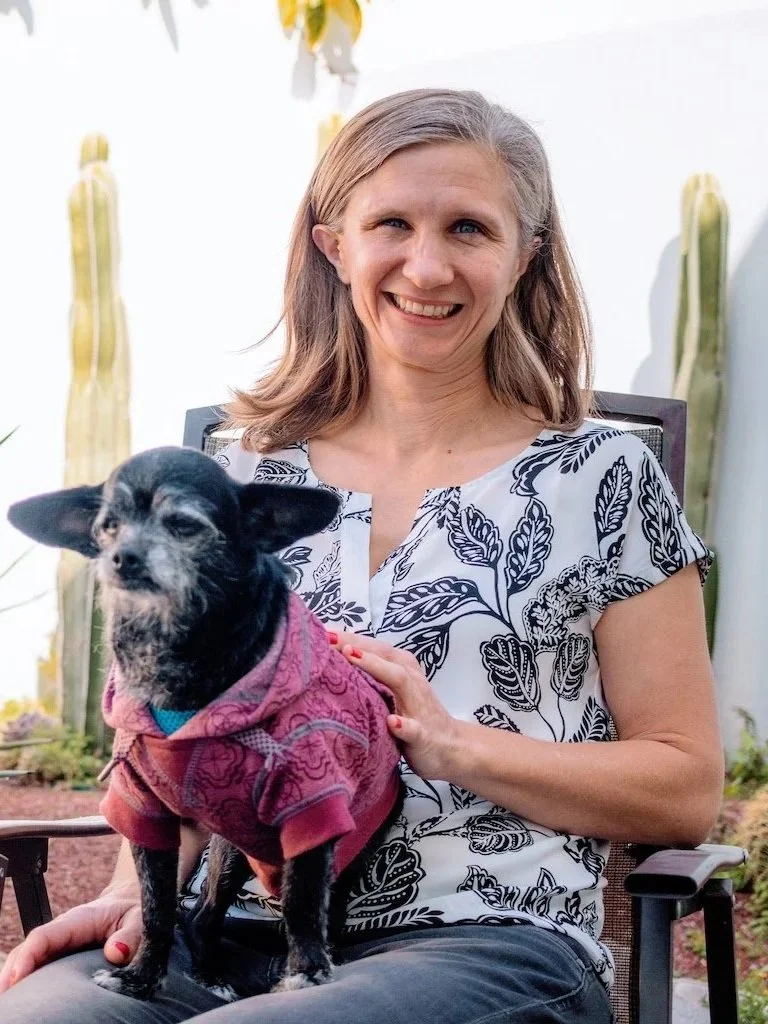 A woman with shoulder-length blonde hair and a patterned blouse smiling while sitting in a chair outdoors, holding a small dog dressed in a pink hoodie.