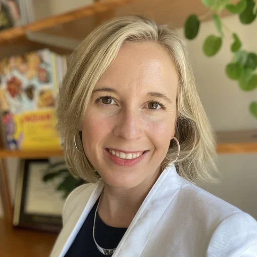 A woman with shoulder-length blond hair, smiling in a home or office setting, wearing a white blazer, hoop earrings, and a black top. There are shelves with books and a green plant in the background.