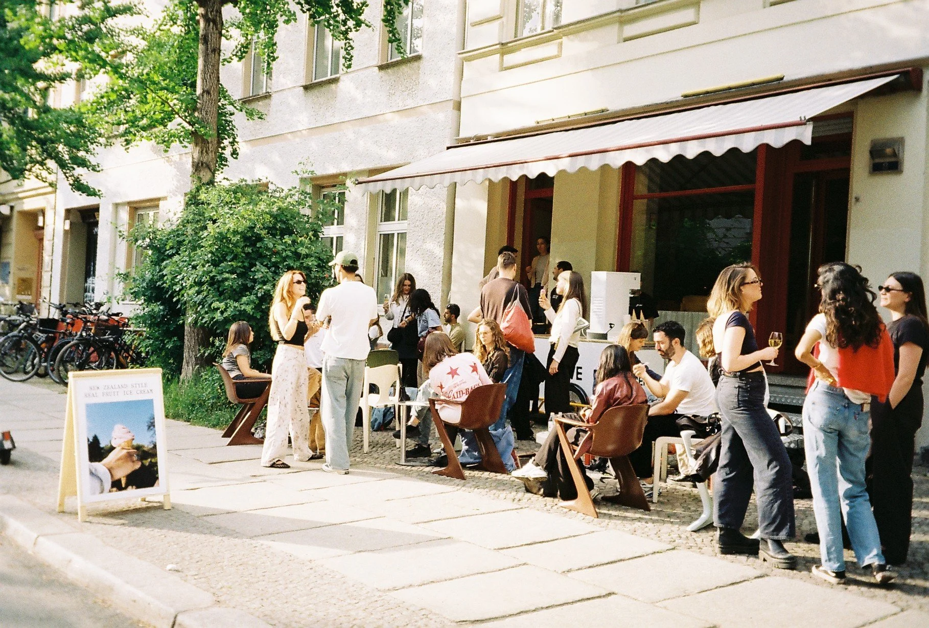 People socializing outside a cafe on a sunny day, with some standing and others sitting at tables, with bicycles parked nearby and a sign advertising ice cream.