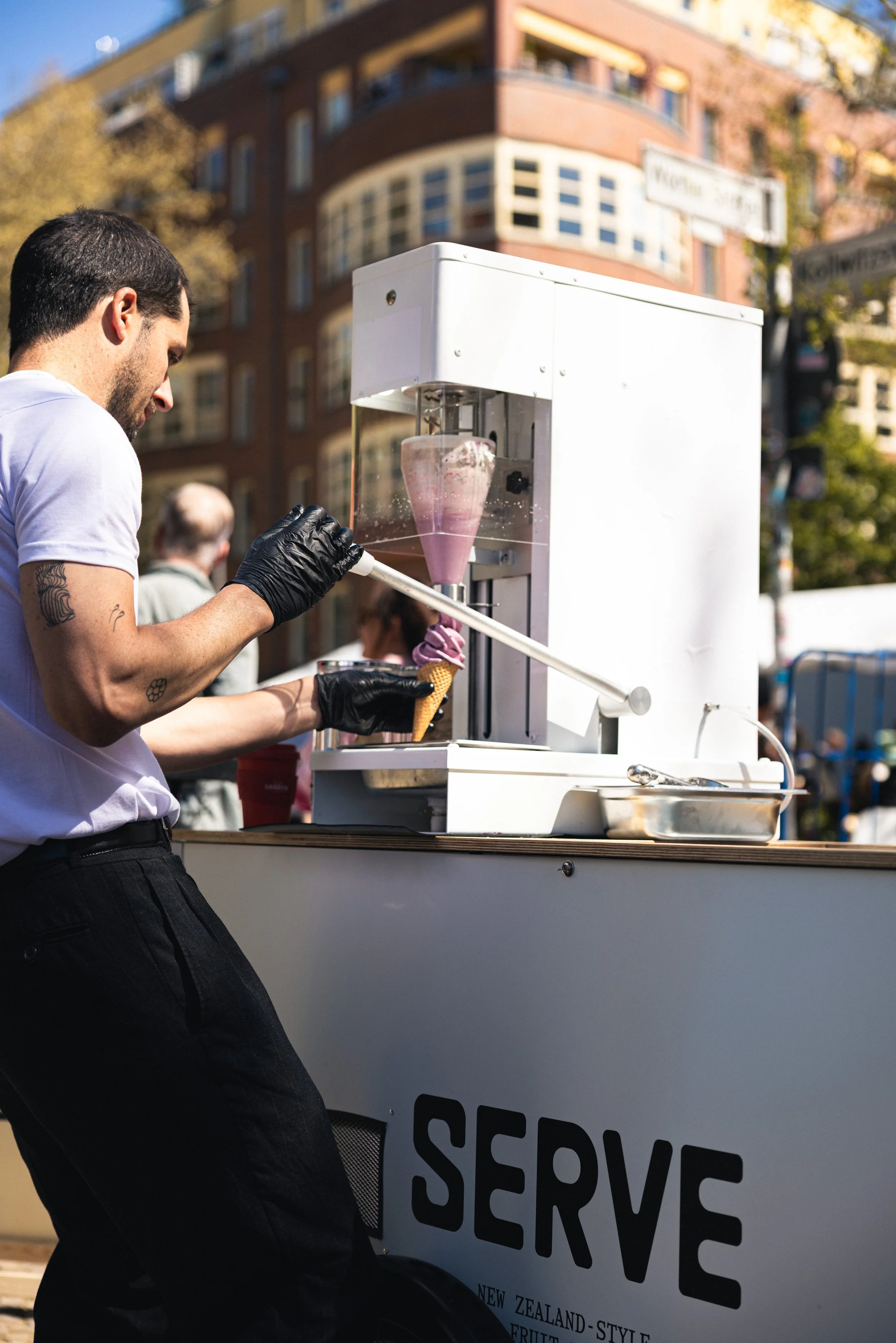 A man in a white shirt and black gloves serving ice cream from an outdoor cart with a sign that says 'We Serve' in a city with buildings and trees in the background.