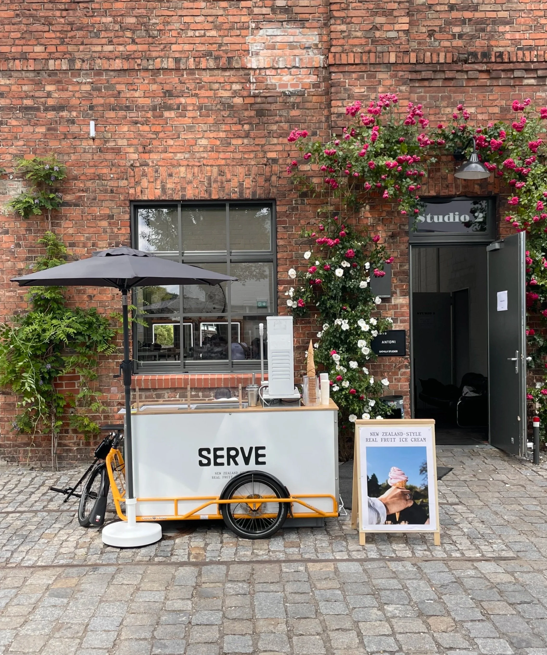 A mobile ice cream cart positioned on a cobblestone street outside a brick building, decorated with blooming pink and white roses. The cart features a bicycle wheel and a canopy, and has a sign that reads 'SERVE.' An umbrella is partially visible above the cart, and a framed picture of an ice cream cone is placed in front of the entrance to the building, which has a sign indicating it is a studio.