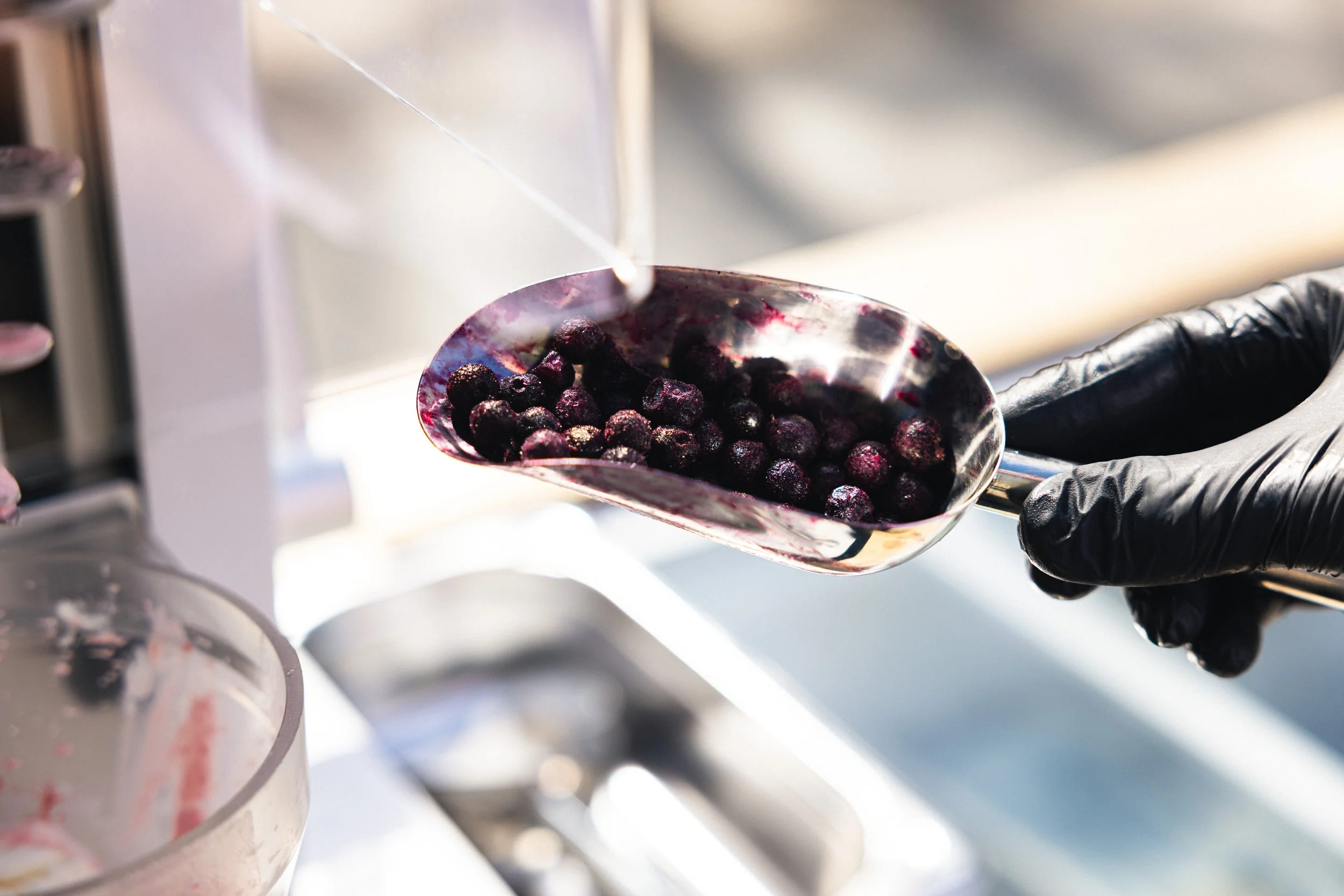 A close-up of a hand wearing a black glove holding a silver scoop filled with purple blueberries in a commercial setting.