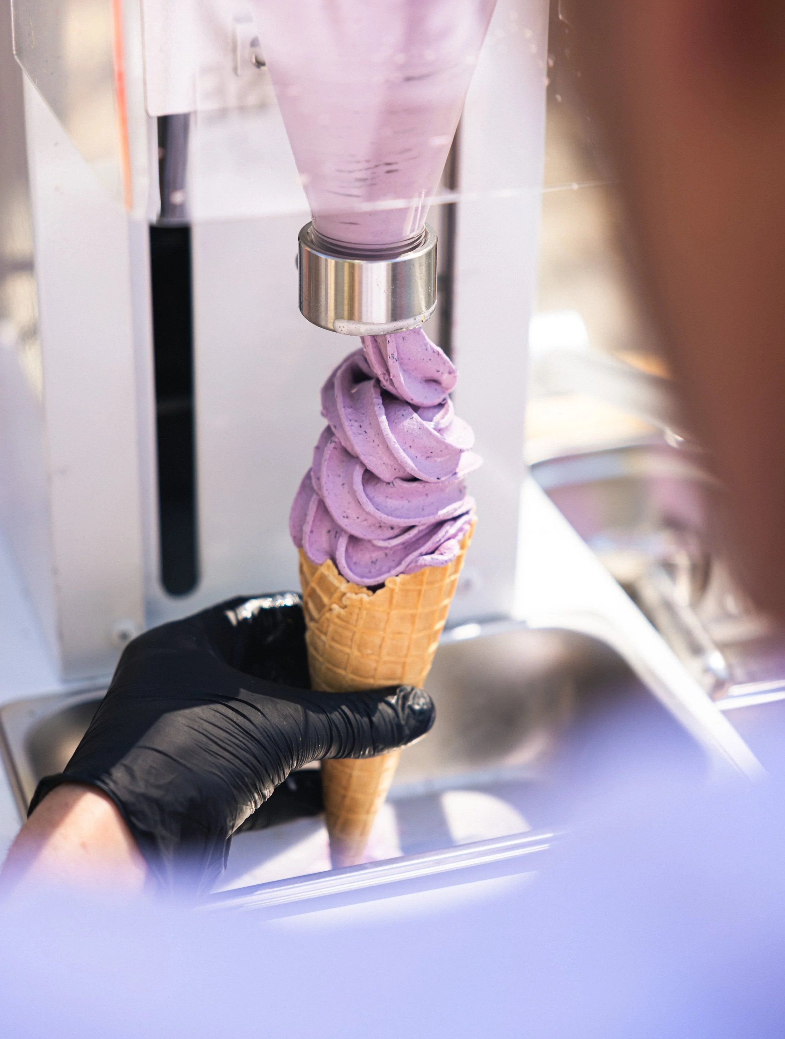 A person wearing a black glove is holding a purple soft serve ice cream cone under a machine dispensing the ice cream.