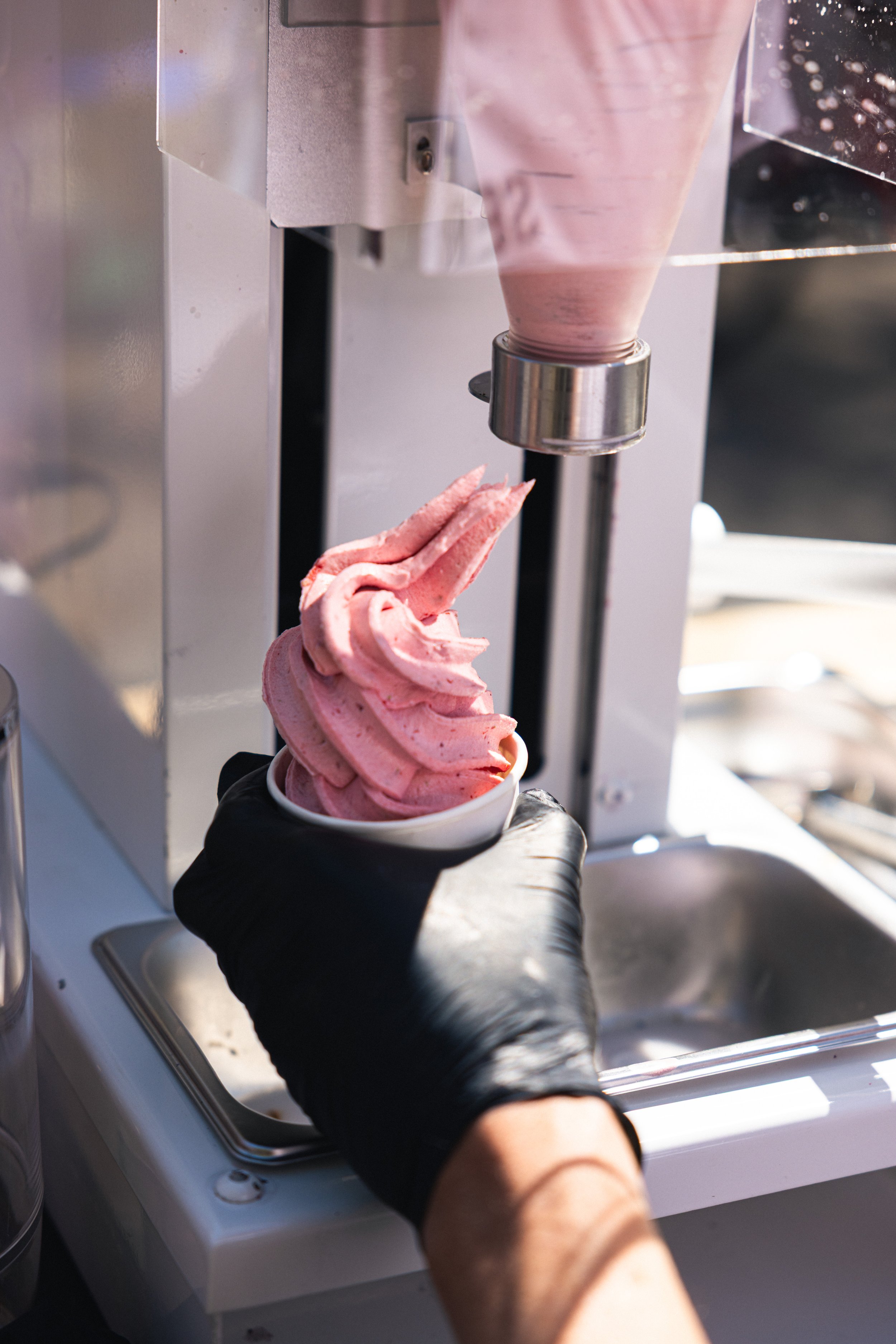 Person wearing black gloves serves pink soft serve ice cream from a machine into a white cup, outdoors with natural sunlight.