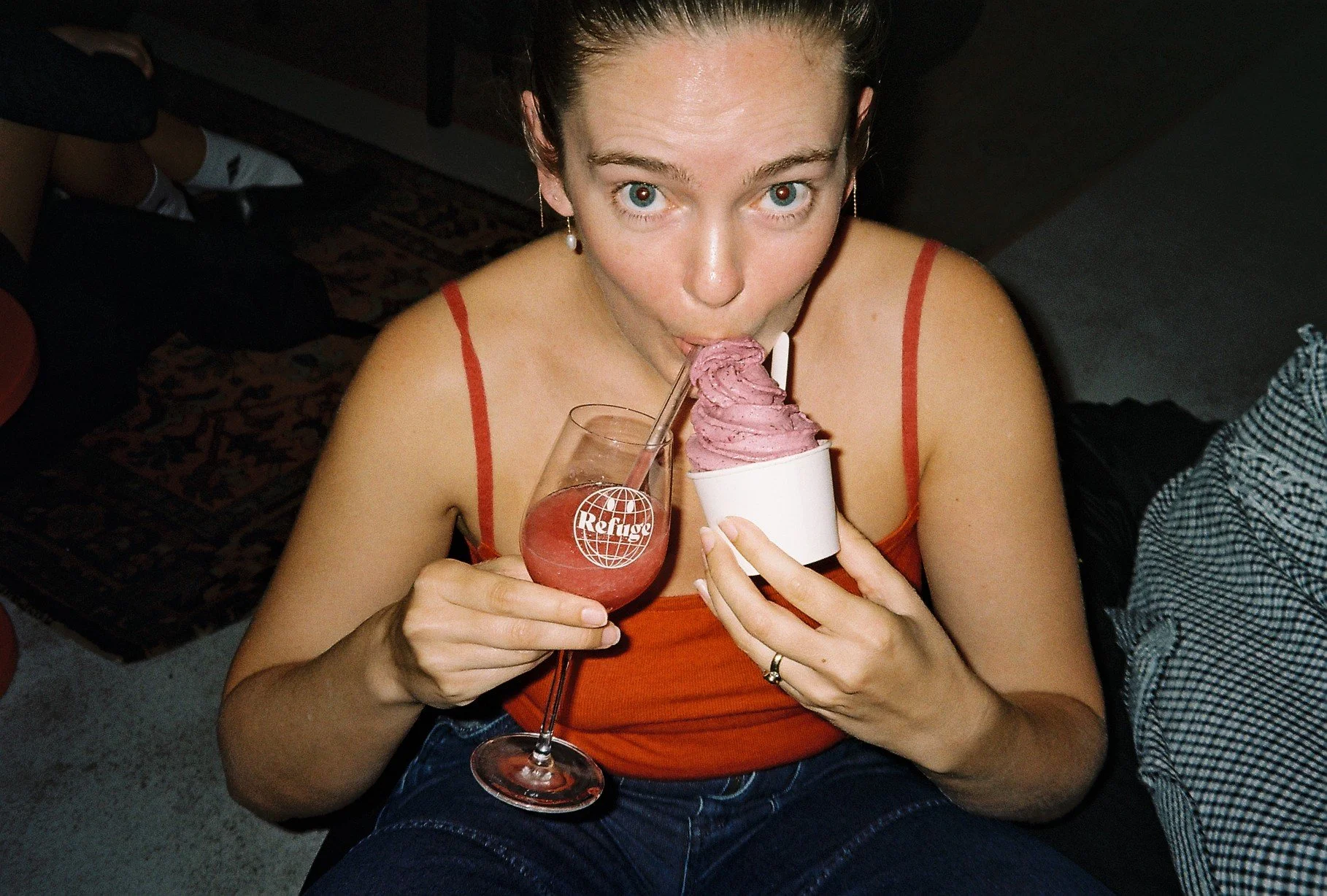 A woman with blue eyes and earrings is holding a glass of pink drink with the logo 'Refuge' and a straw, and a cup of purple soft-serve ice cream with a straw, while looking up at the camera.