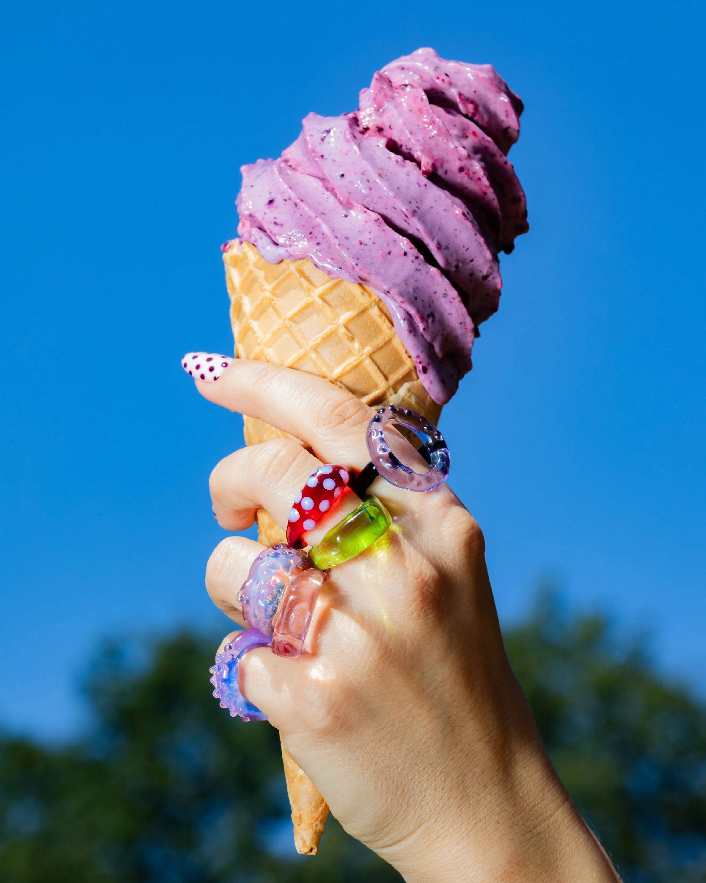 A hand holding a waffle cone with two scoops of purple raspberry ice cream against a blue sky background.