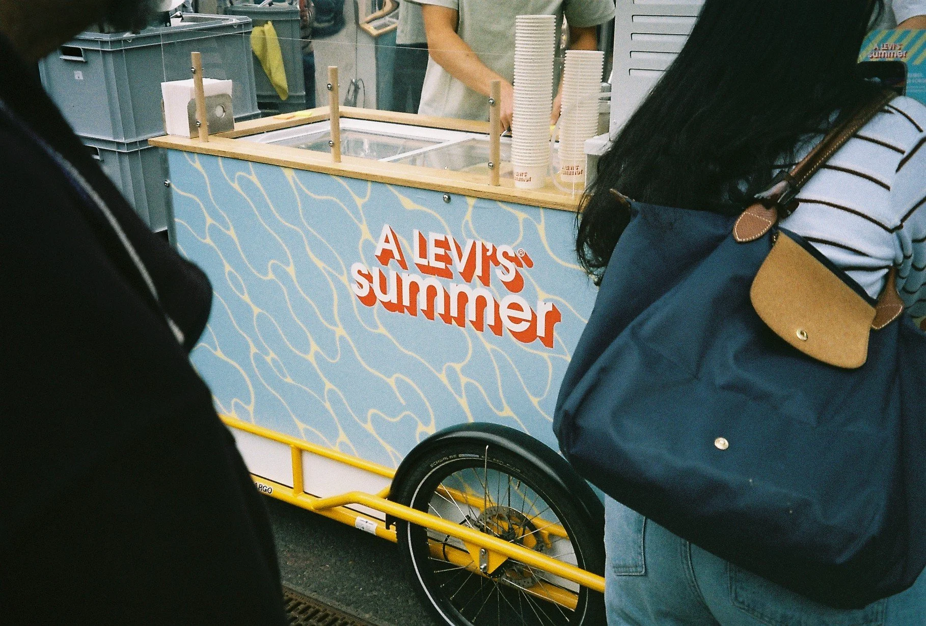 People standing in front of a Levi's Summer ice cream stand, with a bicycle attached to a yellow cart and a person wearing a striped shirt carrying a navy bag.