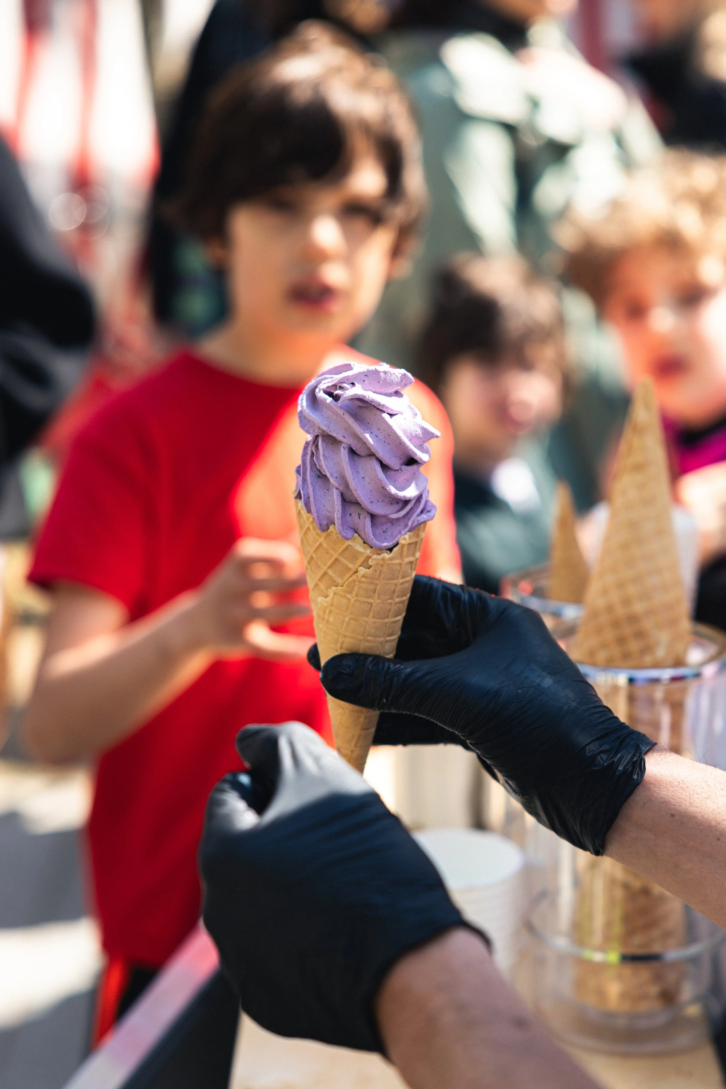 A person wearing black gloves holding a cone of purple ice cream. In the background, there are children, one wearing a red shirt, at an outdoor ice cream stand or festival.