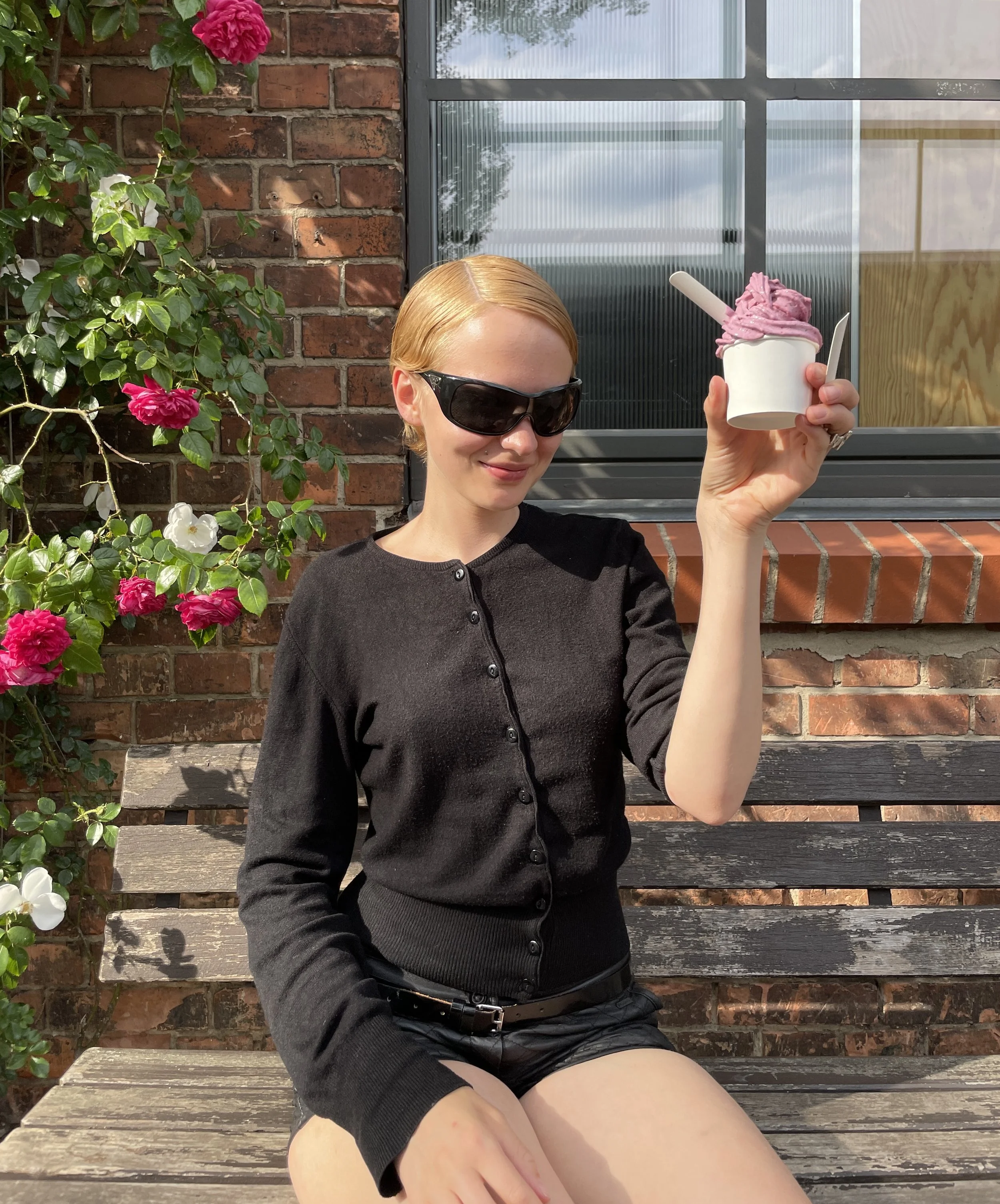 A young woman with blonde hair, wearing black sunglasses, a black cardigan, and black shorts, is sitting on a wooden bench outside near a brick wall and window, holding a cup of pink soft serve ice cream in her right hand.