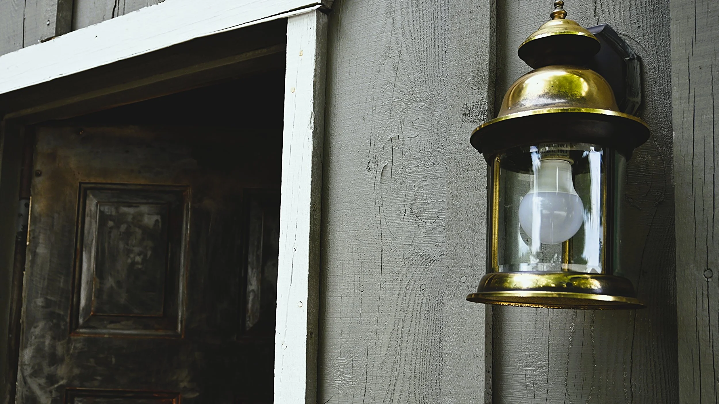 A brass and glass lantern with a white light bulb inside, mounted on a wooden wall outside a rustic building.