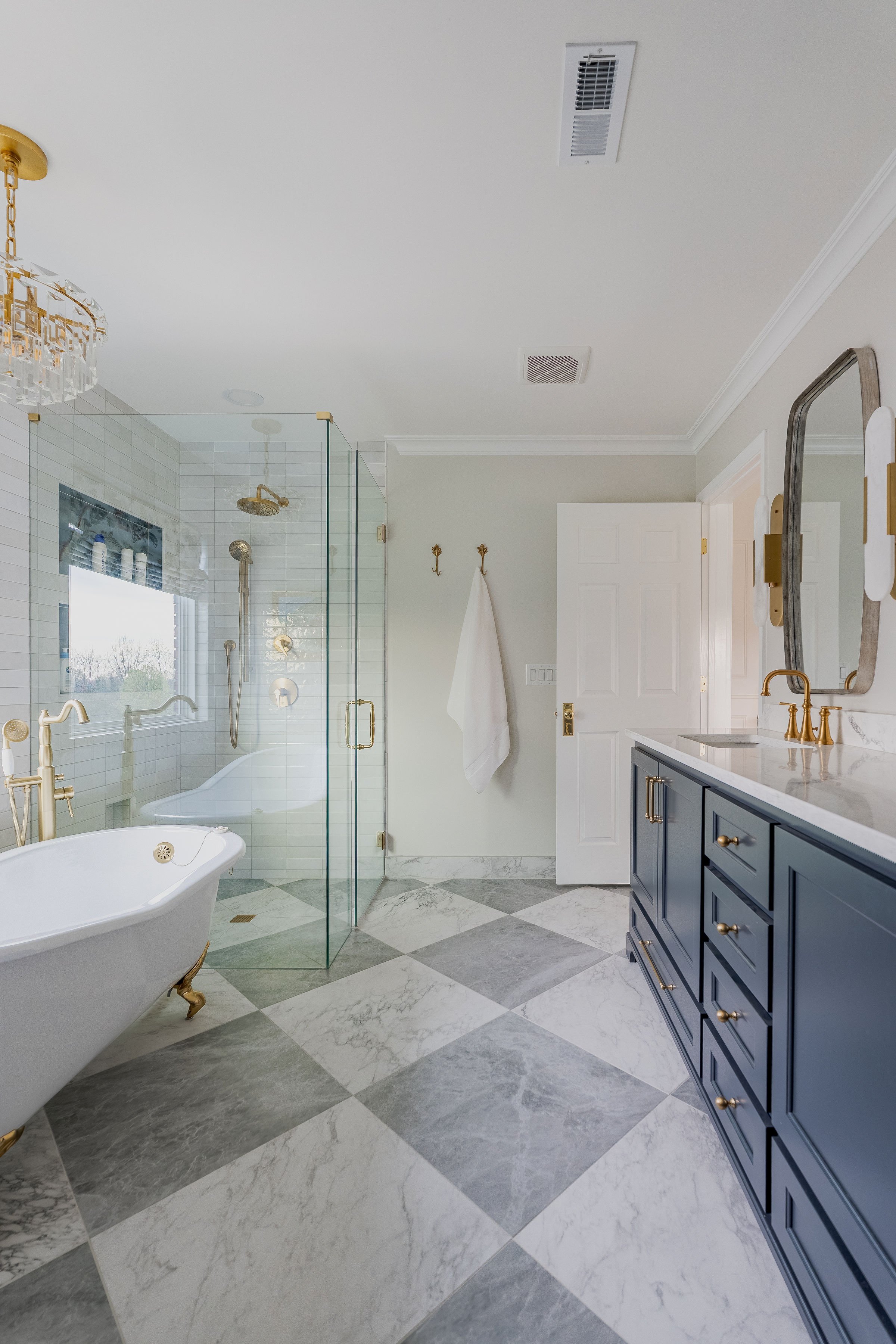 A bathroom with a freestanding clawfoot bathtub, a glass-enclosed shower with gold fixtures, a blue vanity with gold knobs, a large mirror, marble tile flooring, and a white ceiling with HVAC vents.
