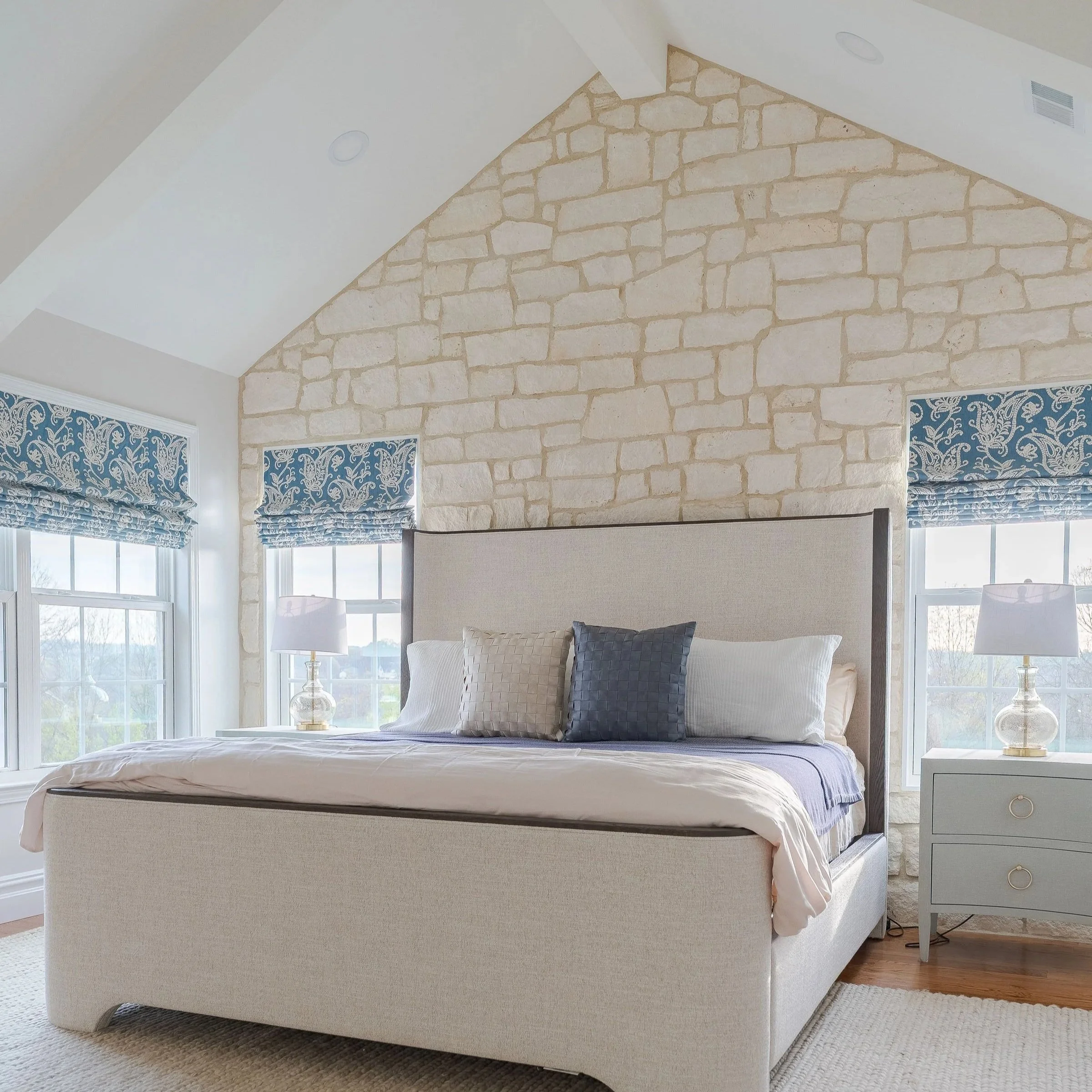 A cozy bedroom with a large bed featuring beige and black pillows, a white duvet, a light-colored upholstered bed frame, and nightstands with glass lamps, set against a background of a stone accent wall and windows with blue patterned curtains.
