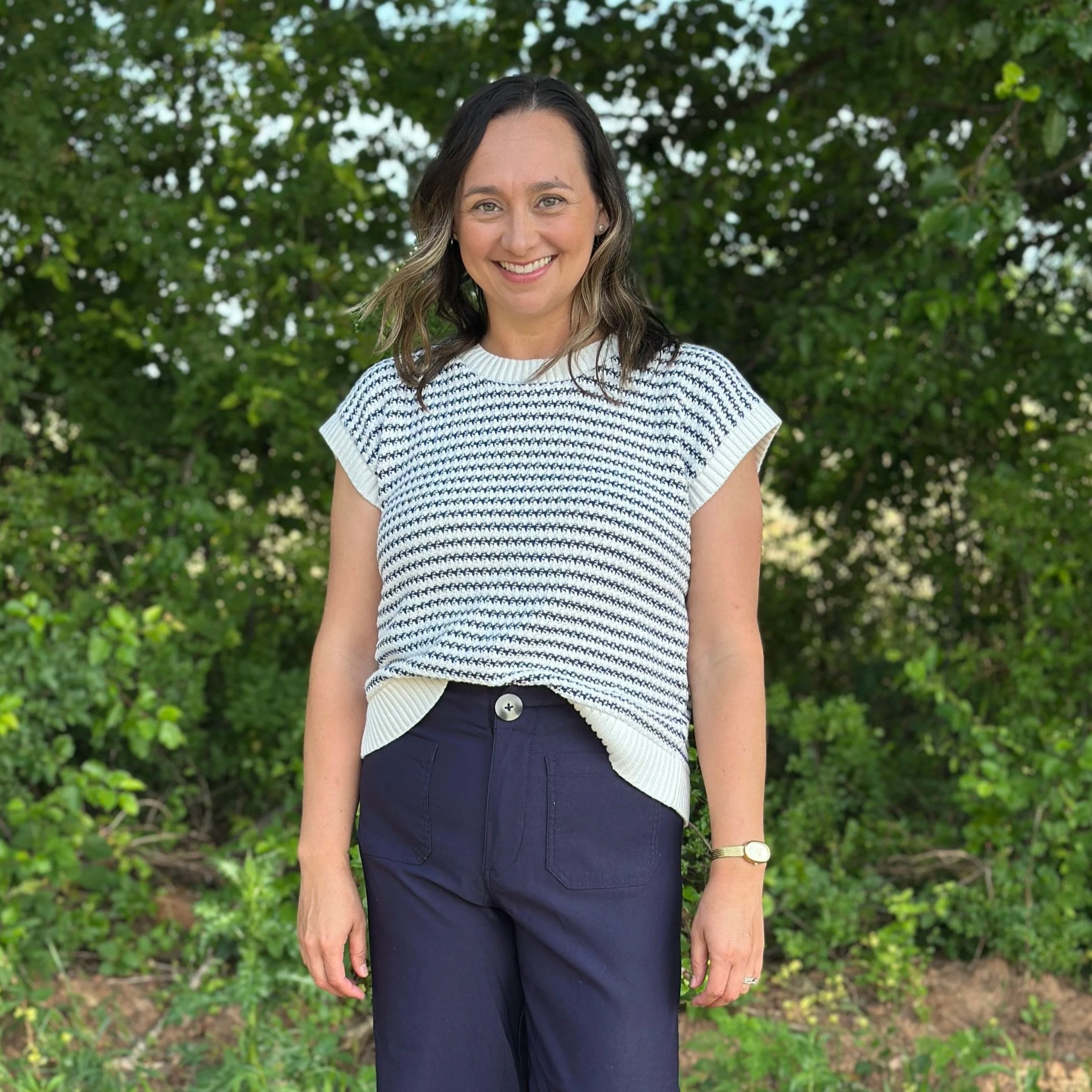 Portrait of a smiling woman with shoulder-length dark hair, wearing a white and navy striped knit top, dark navy pants, and a gold watch on her left wrist, standing outdoors with green foliage in the background.