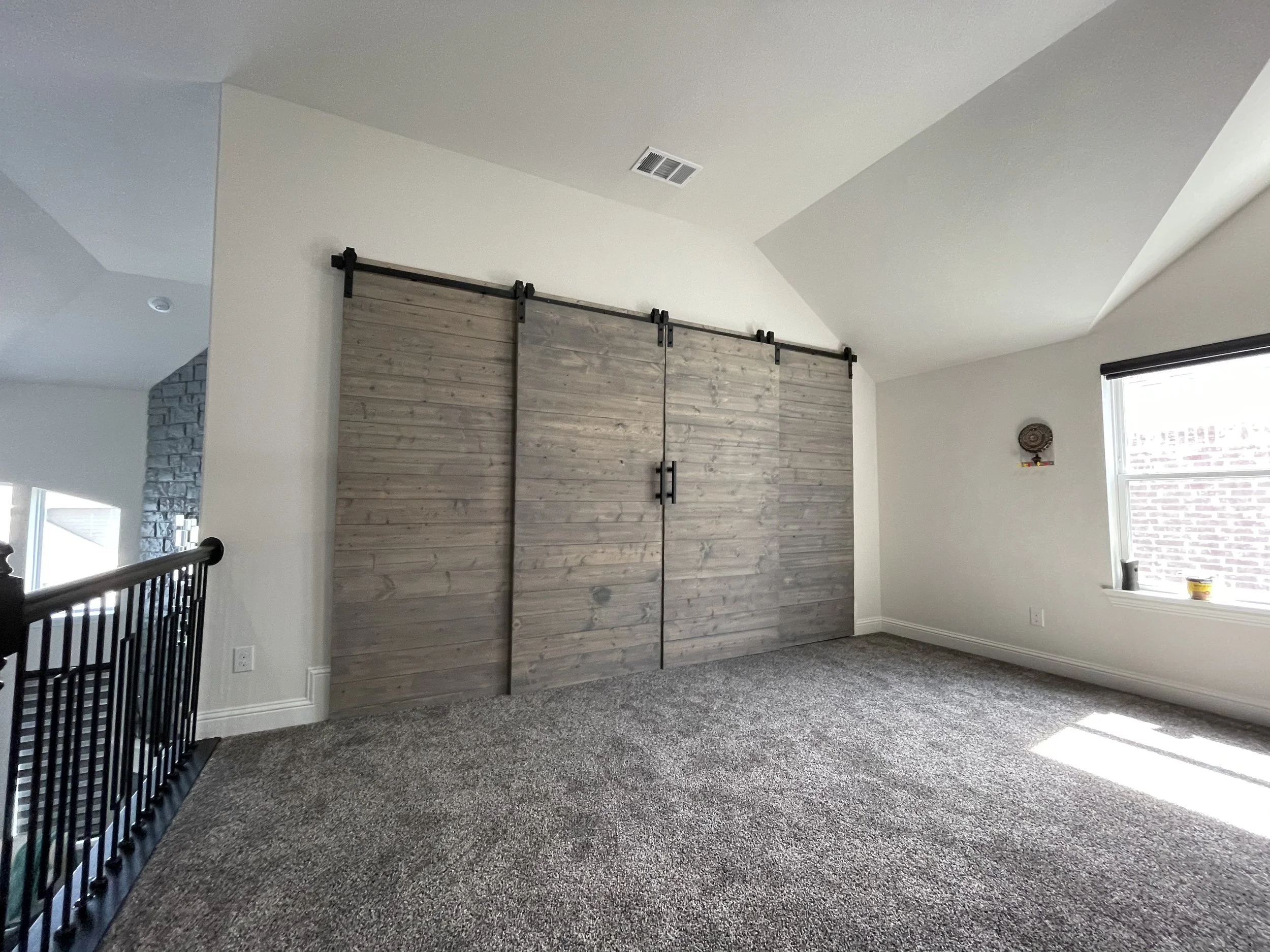 Interior of a room with a high ceiling, featuring a sliding barn door made of wood with black hardware, a barred stair railing, a window with blinds, a small wall decoration, and gray carpet flooring.