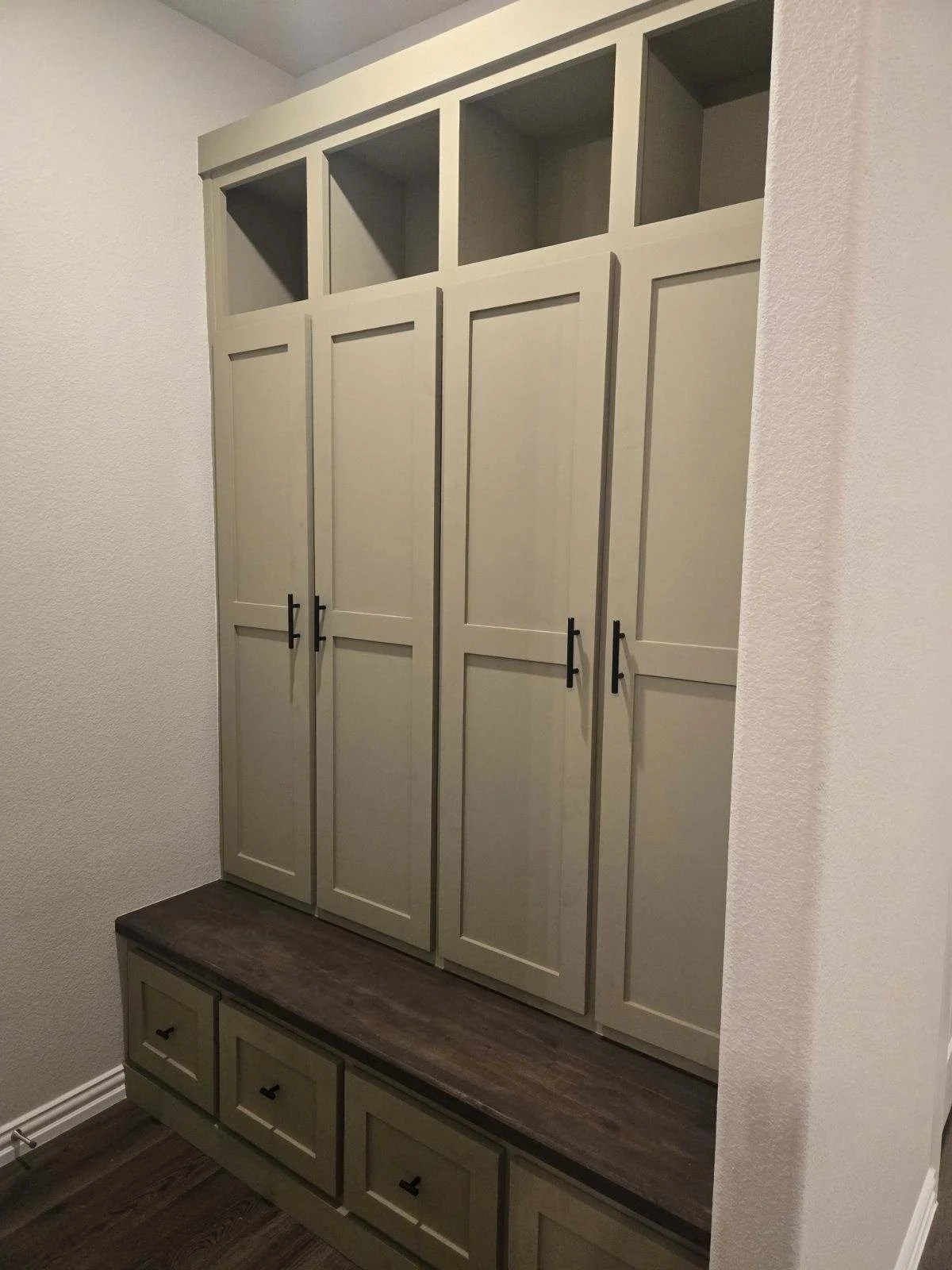 A built-in beige cabinet with black handles, featuring upper open compartments and lower closed doors, with a wooden bench underneath in a room with white textured walls.