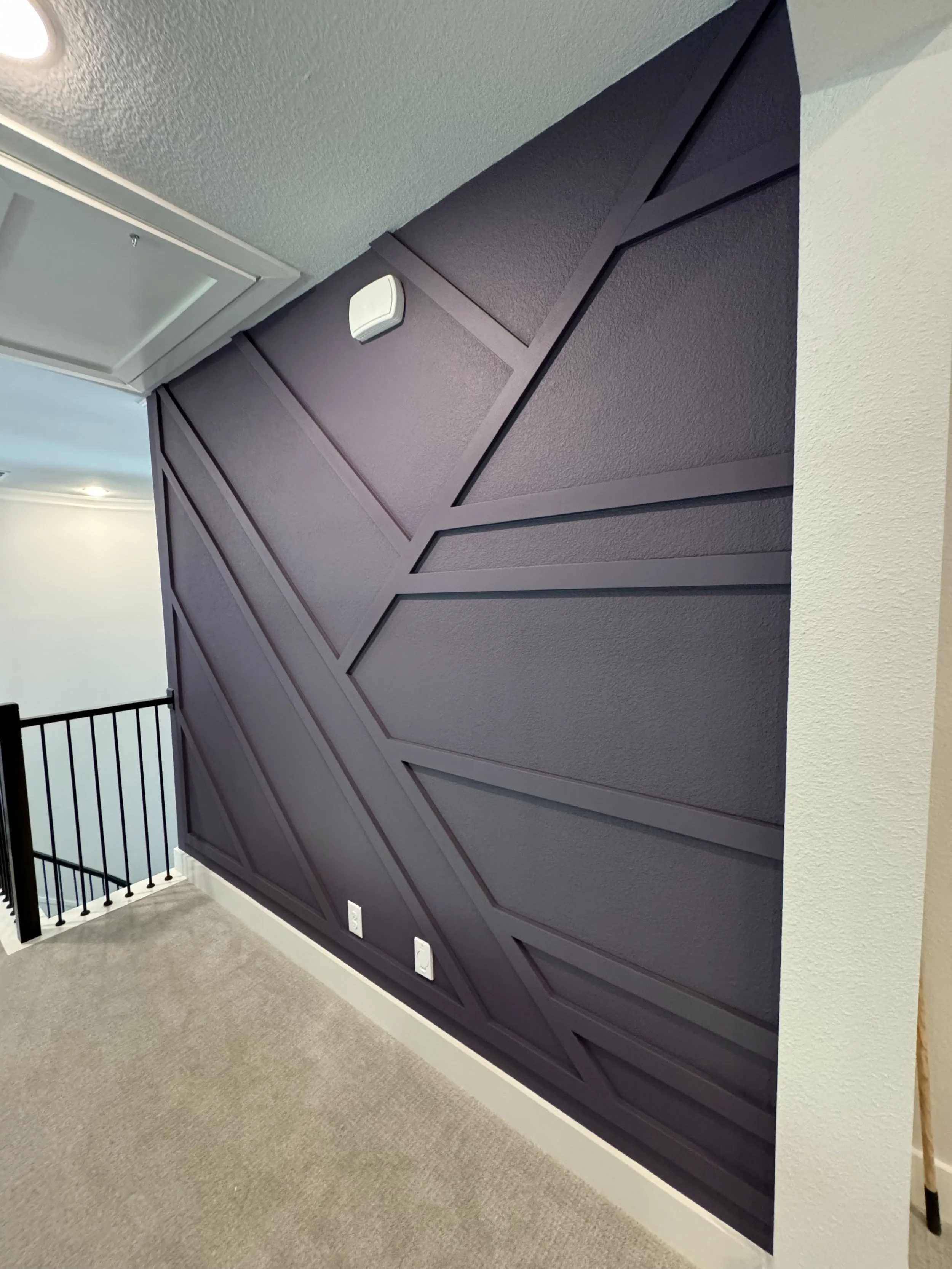 Interior view of a modern room with a dark purple wall featuring geometric paneling, a white vent, three electrical outlets, beige carpet, white trim, and part of a staircase with black railing.