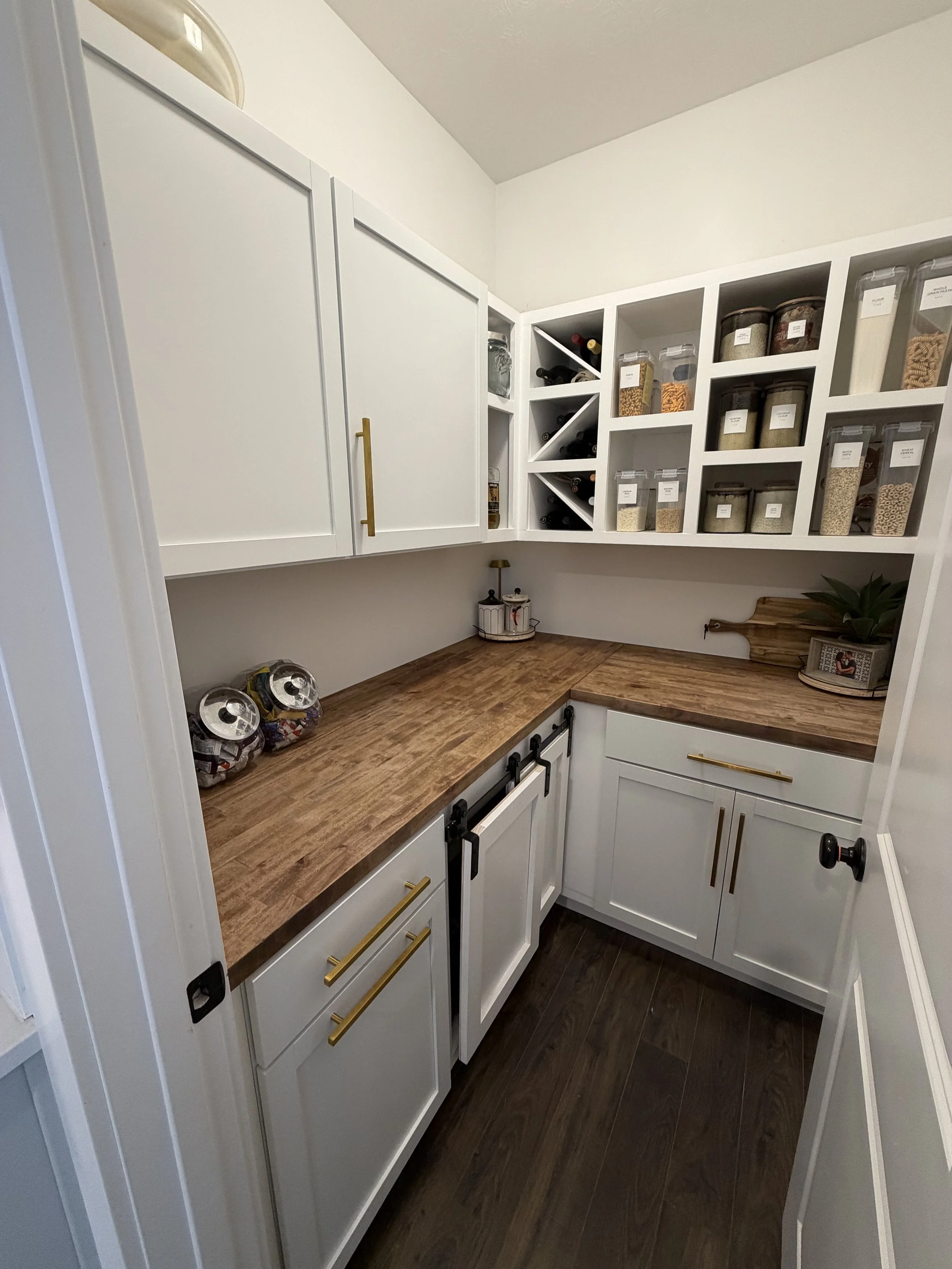 A closet with white cabinets and open shelves holding jars and bottles of spices and ingredients. Wooden countertops with a few decorative items and snacks.