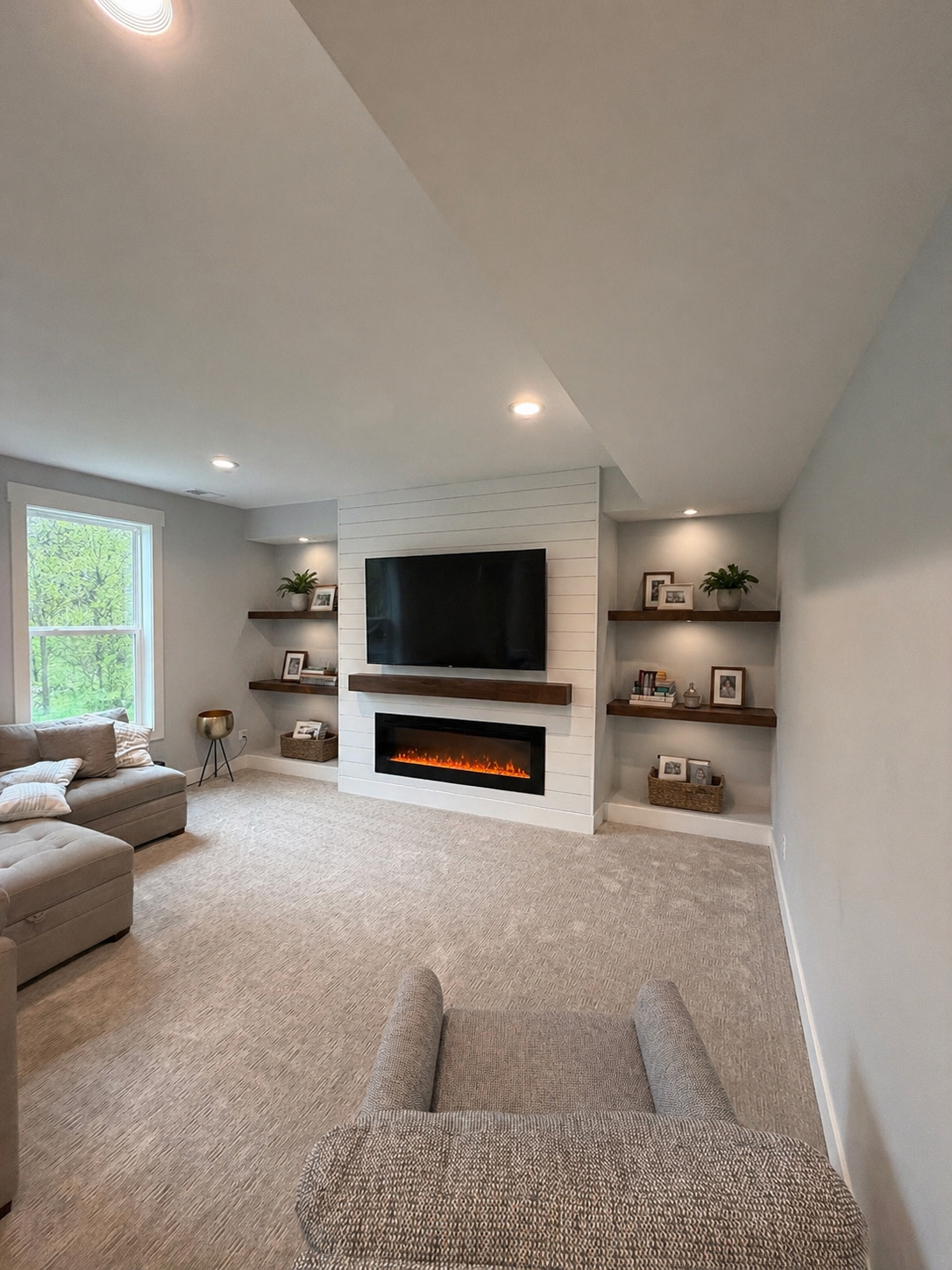Living room with beige carpet, a beige sectional sofa, a window with green trees outside, a white wall with a mounted TV, an electric fireplace below the TV, and floating wooden shelves with decorative items and picture frames.