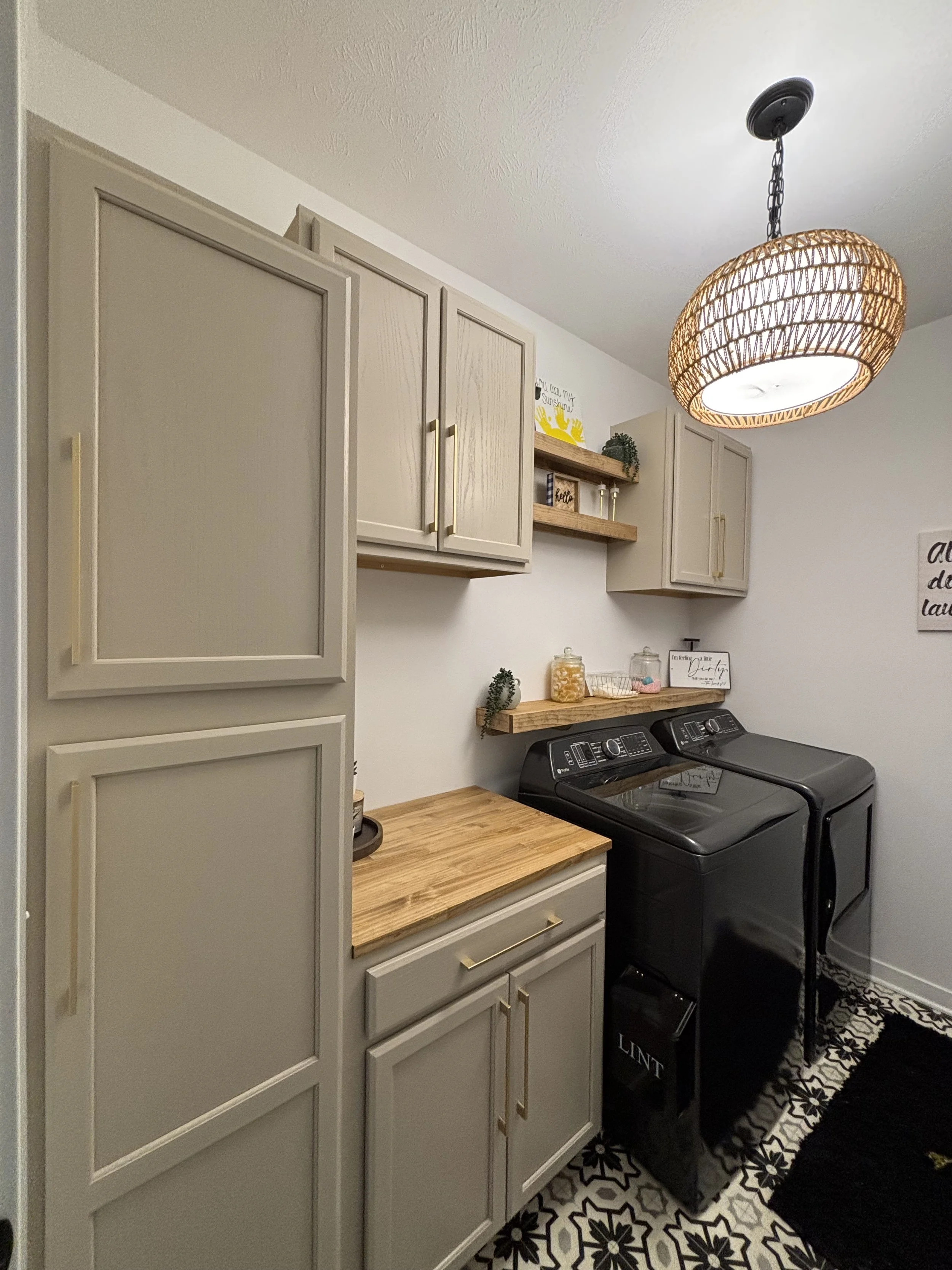 A laundry room with beige cabinets, a wooden countertop, and a black washer and dryer. A wooden shelf holds decorative items and small plants. The room has a black-and-white geometric patterned rug and a rattan pendant light hanging from the ceiling.