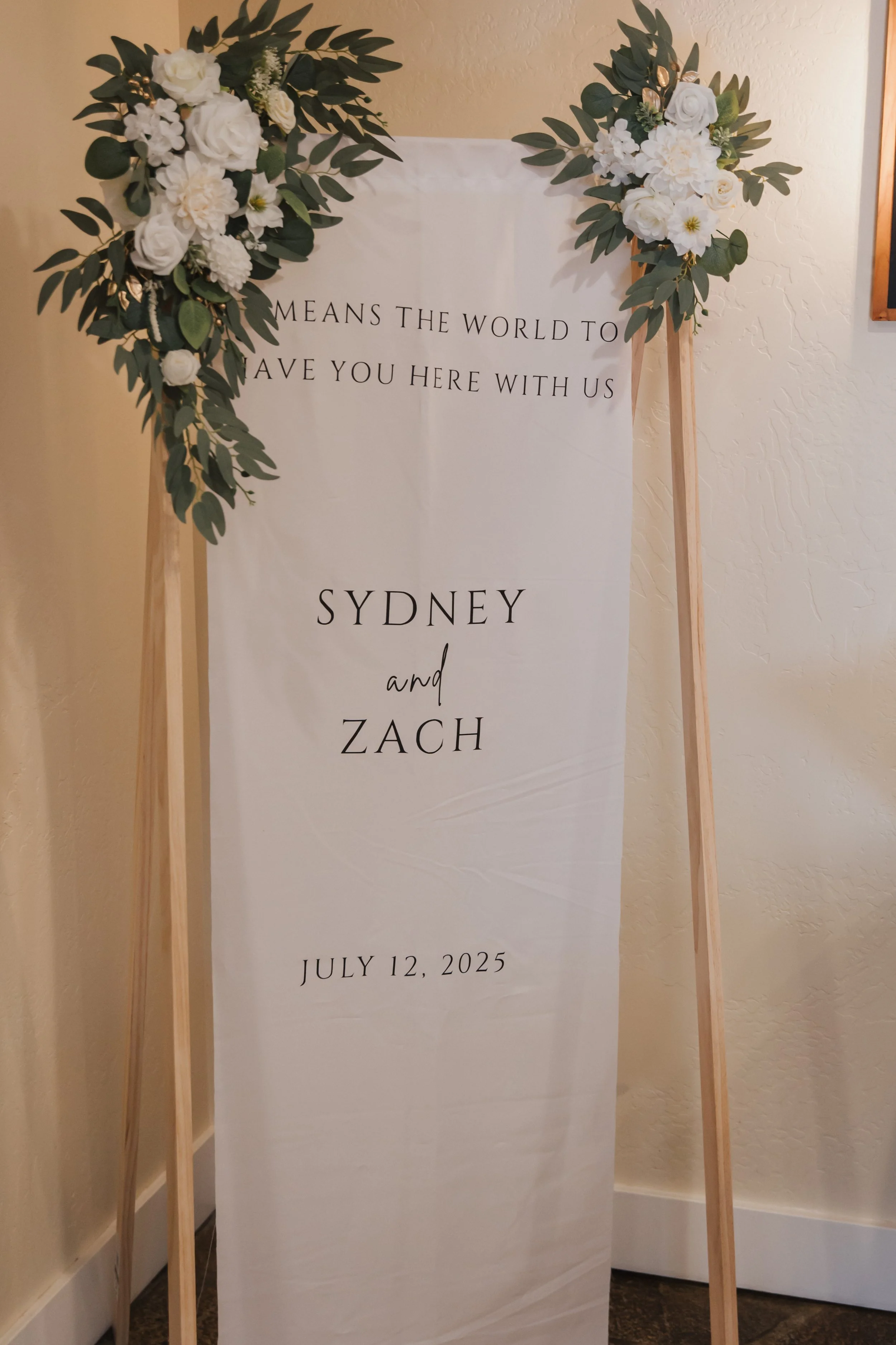 A vertical wedding or celebration sign decorated with white and green floral arrangements, displaying the names Sydney and Zach, and the date July 12, 2025.