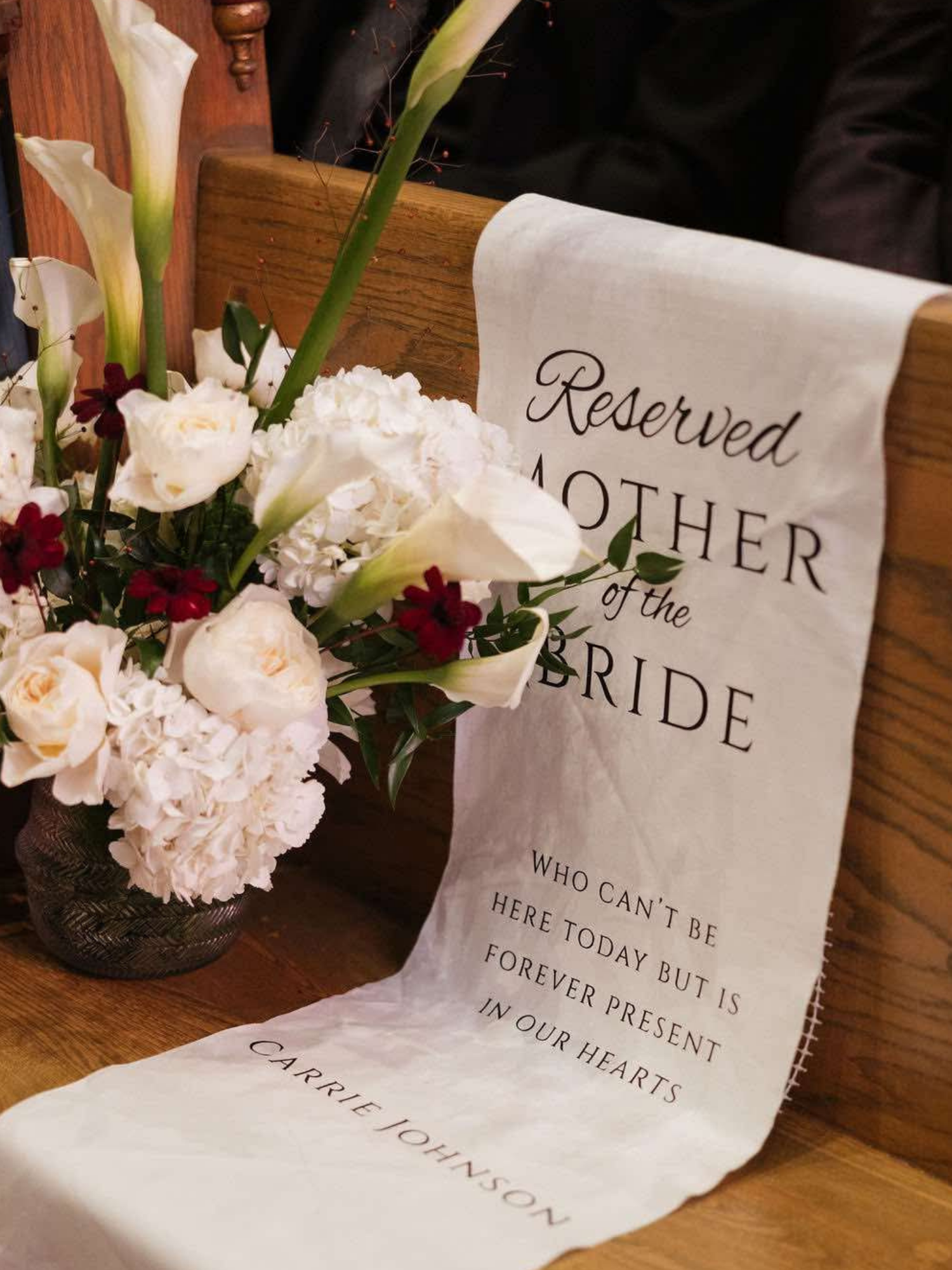 A bouquet of white flowers in a dark vase next to a fabric banner with a reserved mother of the bride message, placed on a wooden table.