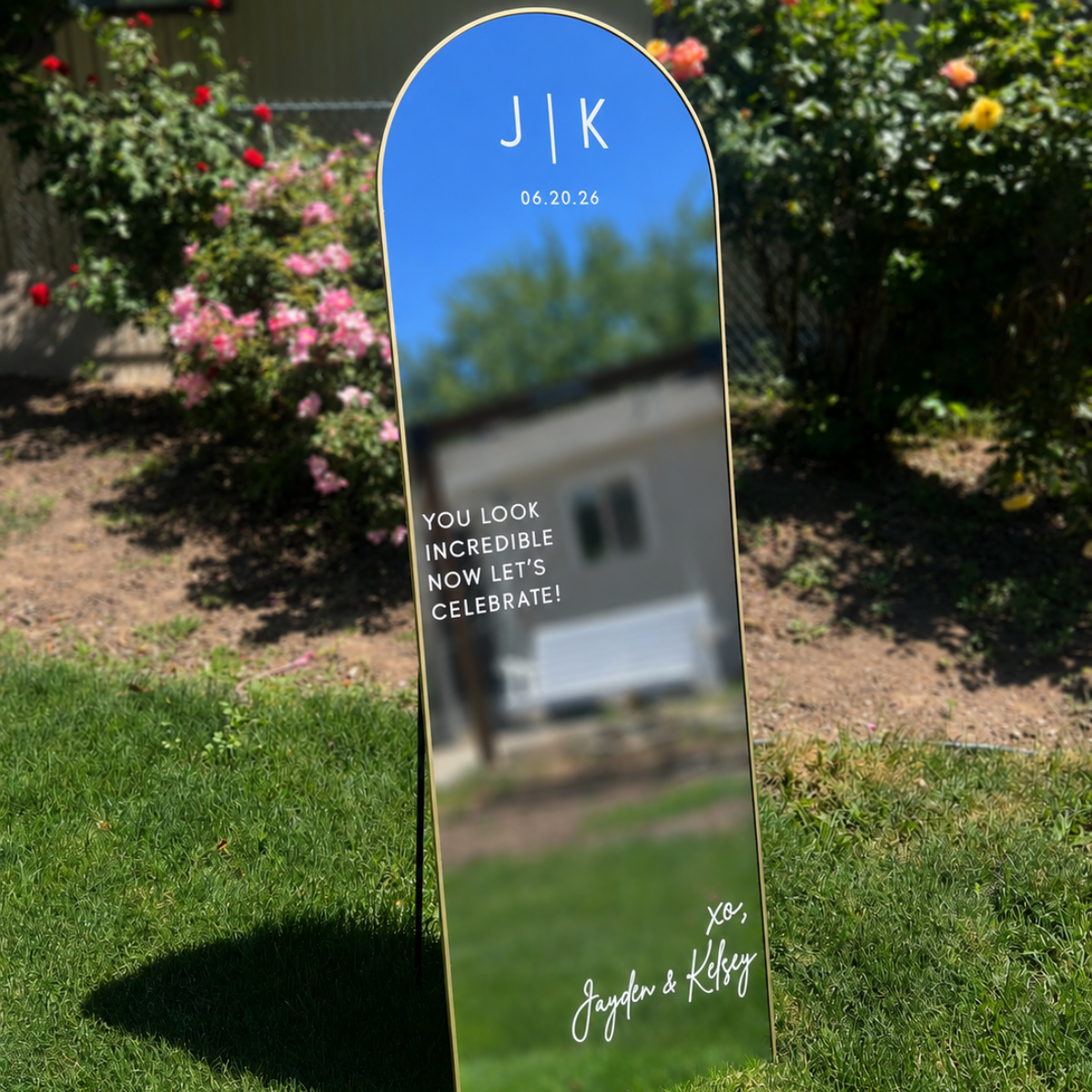 A tall, arched mirror with a gold rim, placed outdoors on the grass, reflecting a blue sky and trees. The mirror has a message for a couple's wedding, including their initials 'J K,' the date 06.20.26, and a congratulatory note: 'You look incredible. Now let's celebrate!' Signed by Jayden and Kelsey.