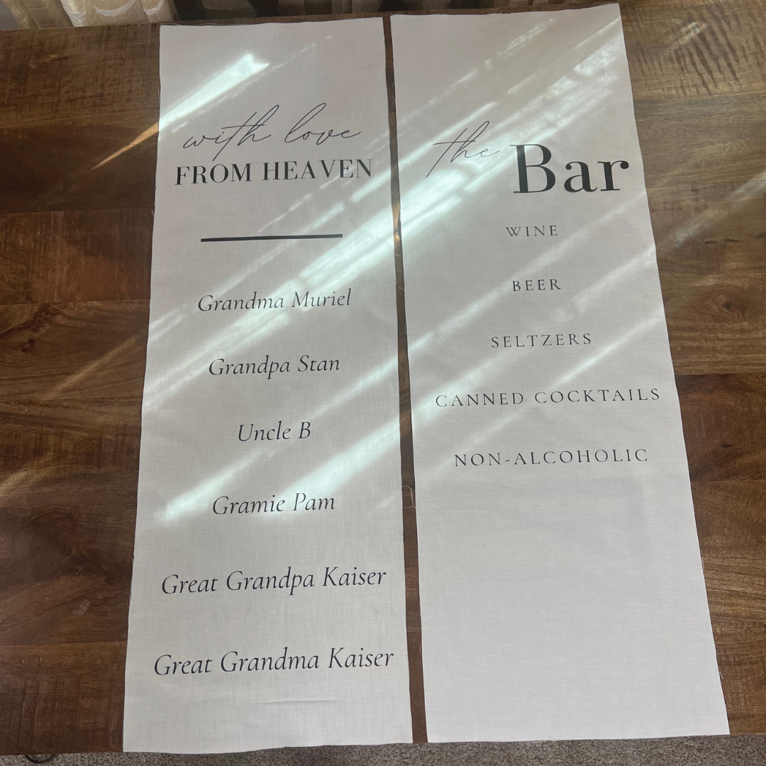 A two-part printed sign on a wooden table. The left part has a message reading "with love FROM HEAVEN" and a list of names: Grandma Muriel, Grandpa Stan, Uncle B, Granie Pam, Great Grandpa Kaiser, Great Grandma Kaiser. The right part lists items available at the bar: wine, beer, seltzers, canned cocktails, non-alcoholic drinks.