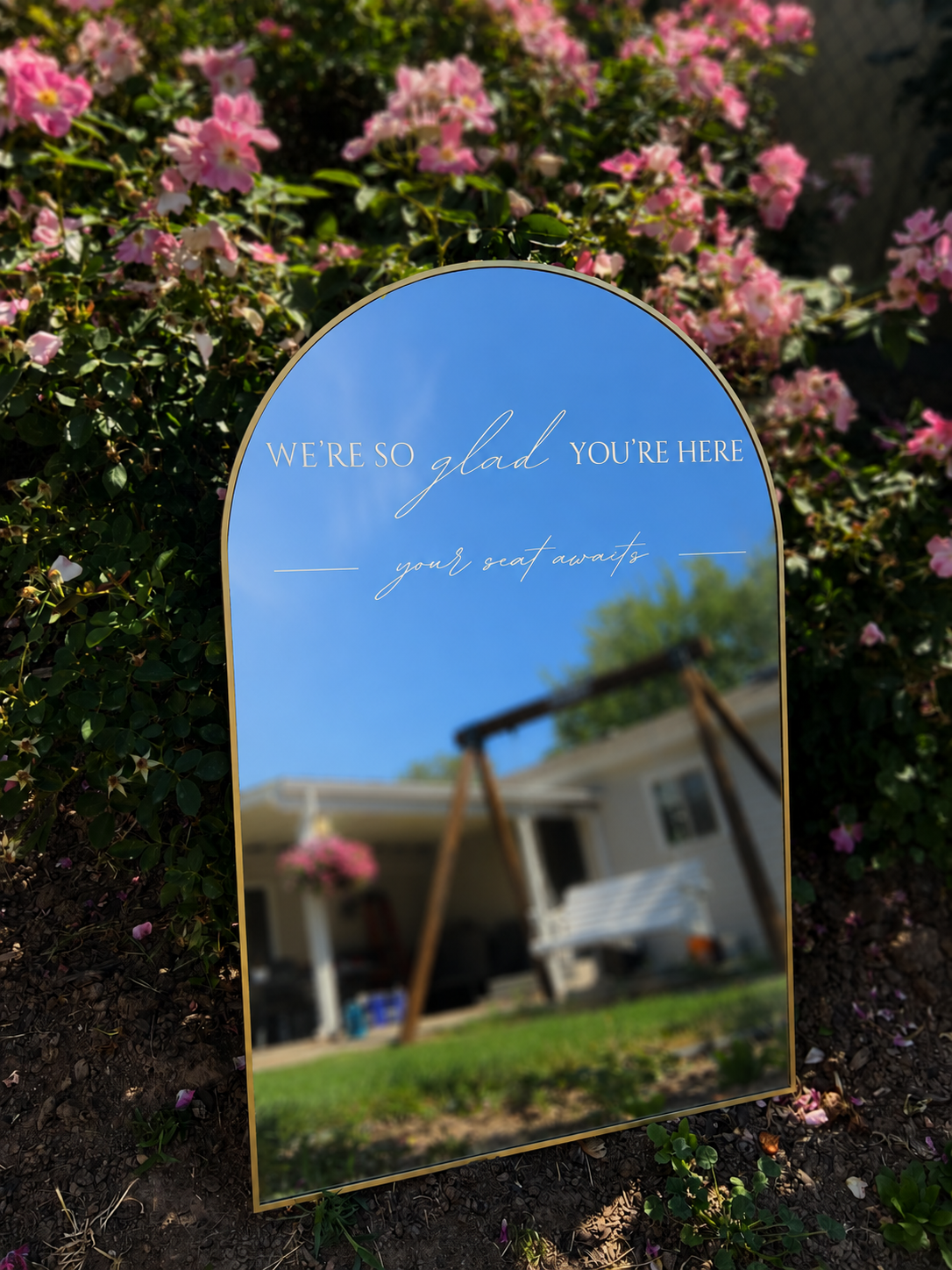 A decorative mirror with a message, placed outdoors among pink flowering bushes, reflects a house with a porch swing and hanging flower basket.