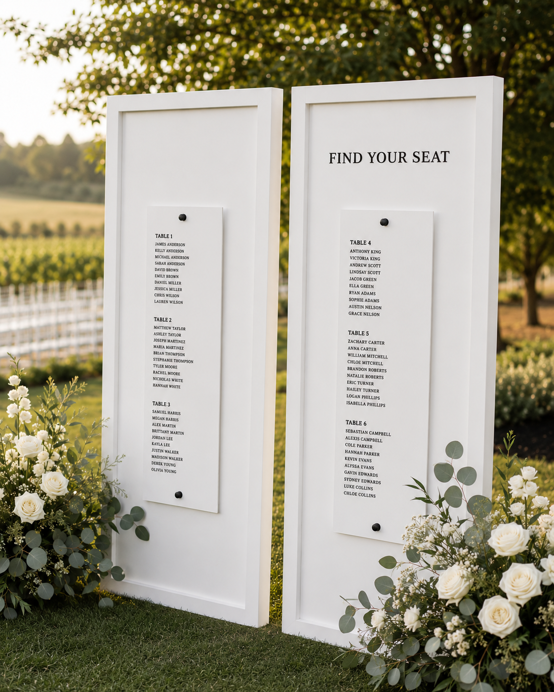 Two white wedding seating chart boards outdoors with black text listing guest names and table numbers, surrounded by white flower arrangements and greenery.