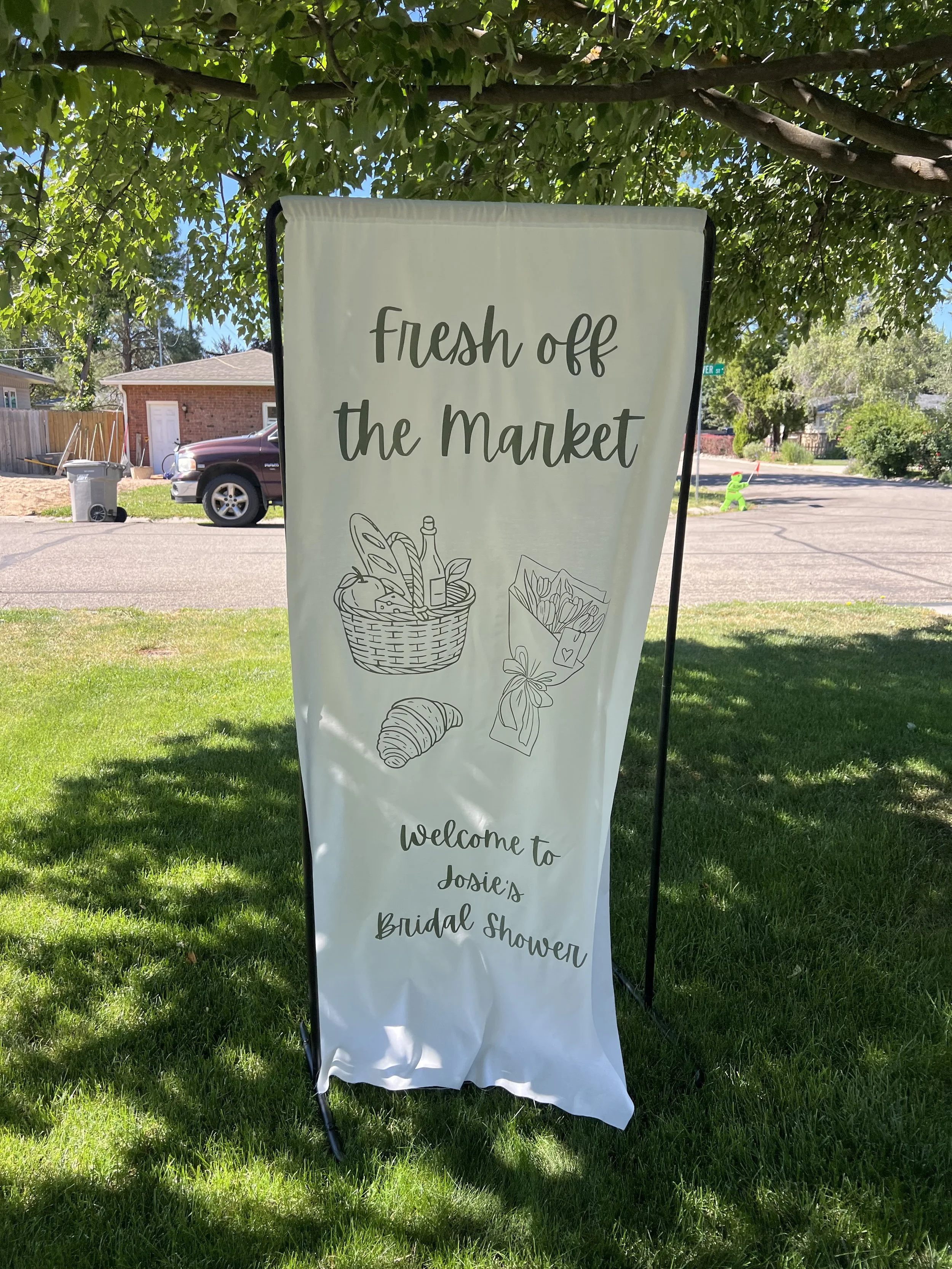 A white banner with black text and illustrations, displaying the words 'fresh off the market' and celebrating Josie's Bridal Shower. The banner is set outdoors under a tree, with a grassy area, a parked car, and residential houses in the background.