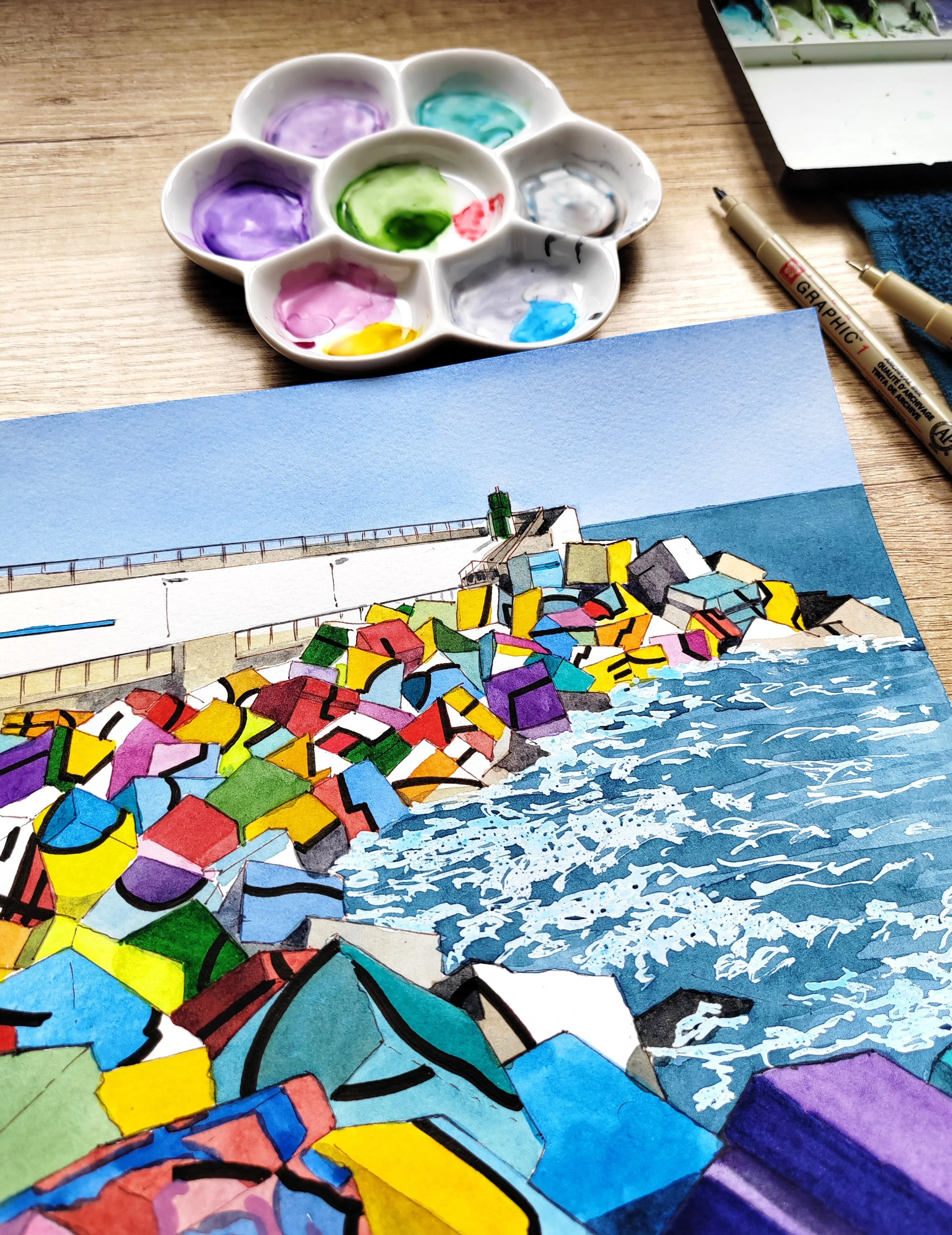Paleta de acuarelas con manchas de pintura de varios colores, y un cuadro de paisaje marino con rocas de colores, playa y mar, al lado de marcadores y una tela azul.