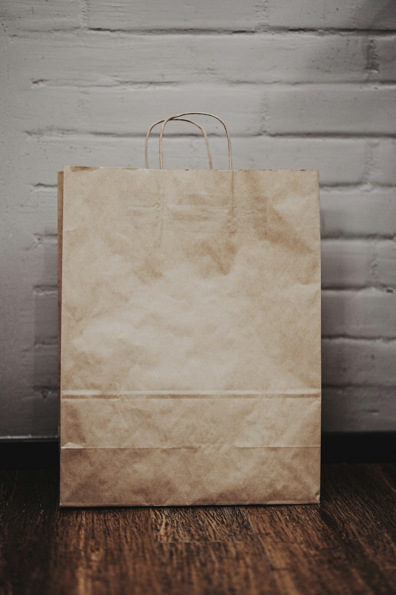 Una bolsa de papel de color marrón claro con asas de cuerda, colocada sobre una superficie de madera de tono oscuro, con una pared de ladrillos pintados de gris en el fondo.